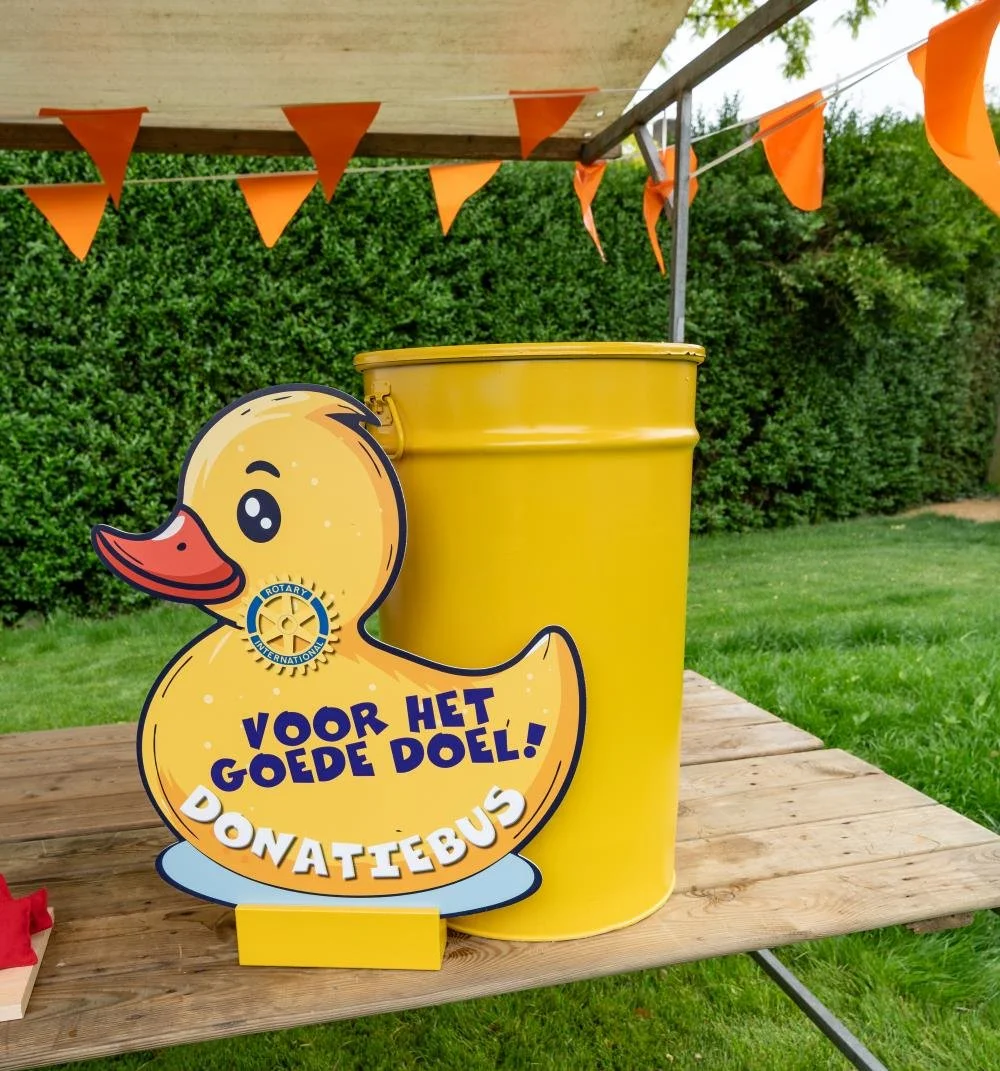 Een gele donatiebak in de vorm van een rubber eend met een bordje dat zegt 'Voor het goede doel! Donatiebus', geplaatst op een houten tafel onder een tent met oranje vlaggetjes, in een groene tuin.
