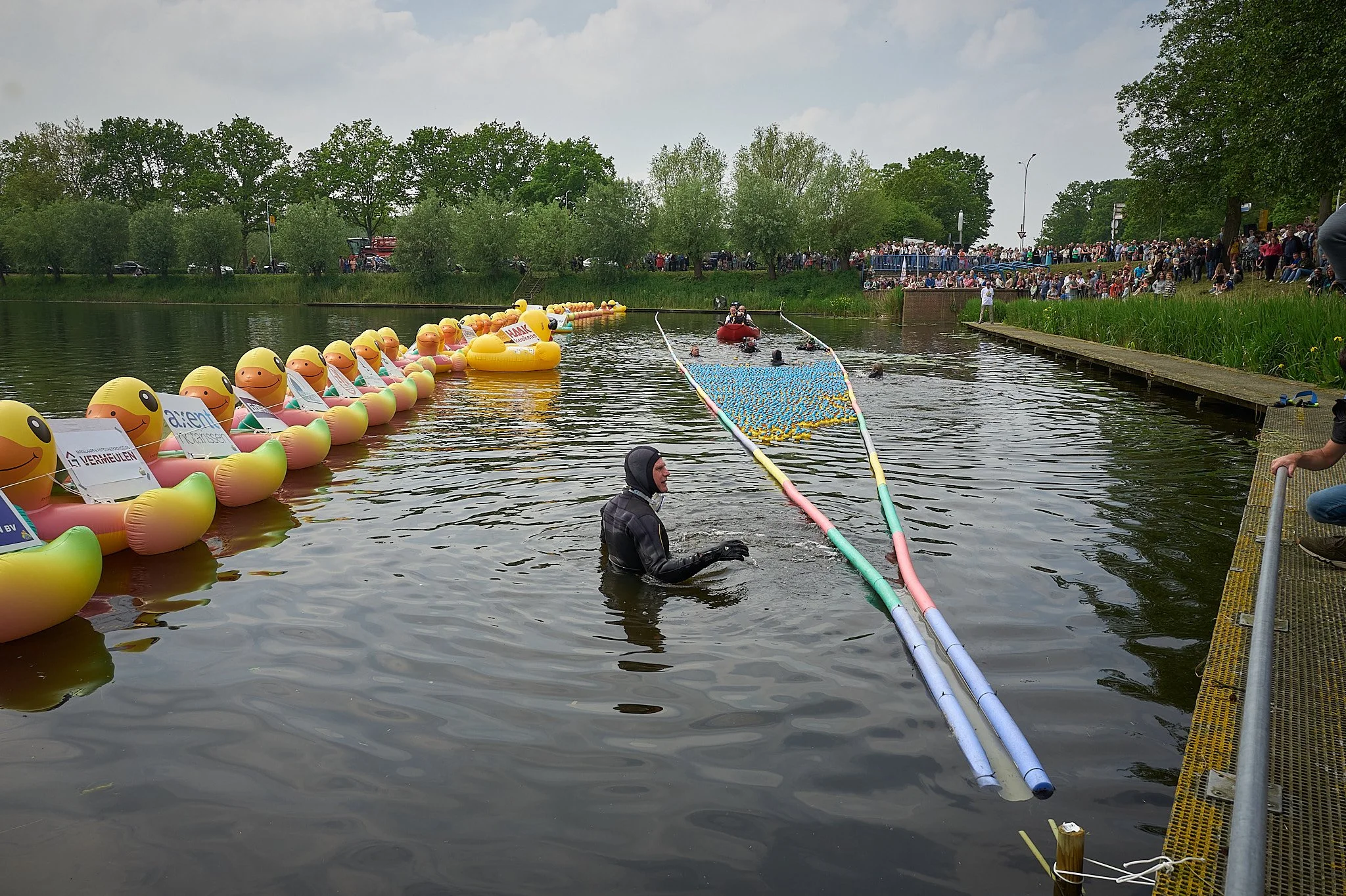 Persoon in water met rubberen ring met smiley gezicht, terwijl er een met andere mensen in een boot op het water is, omlijnd door een menigte in de verte en op de oever.