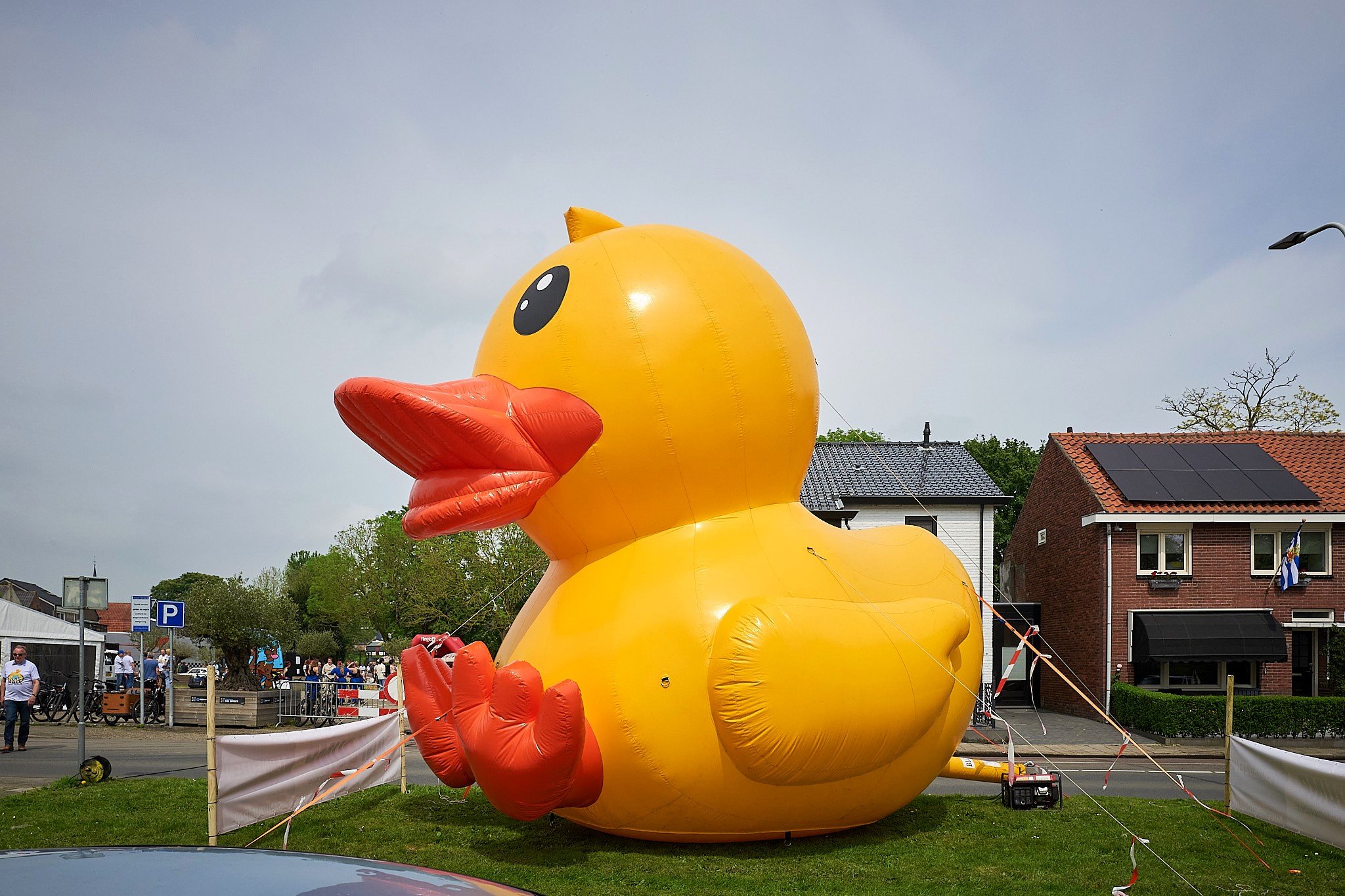 Gigantische gele rubber eend met oranje snavels en voeten op een grasveld, omringd door mensen en gebouwen, mogelijk onderdeel van een evenement.