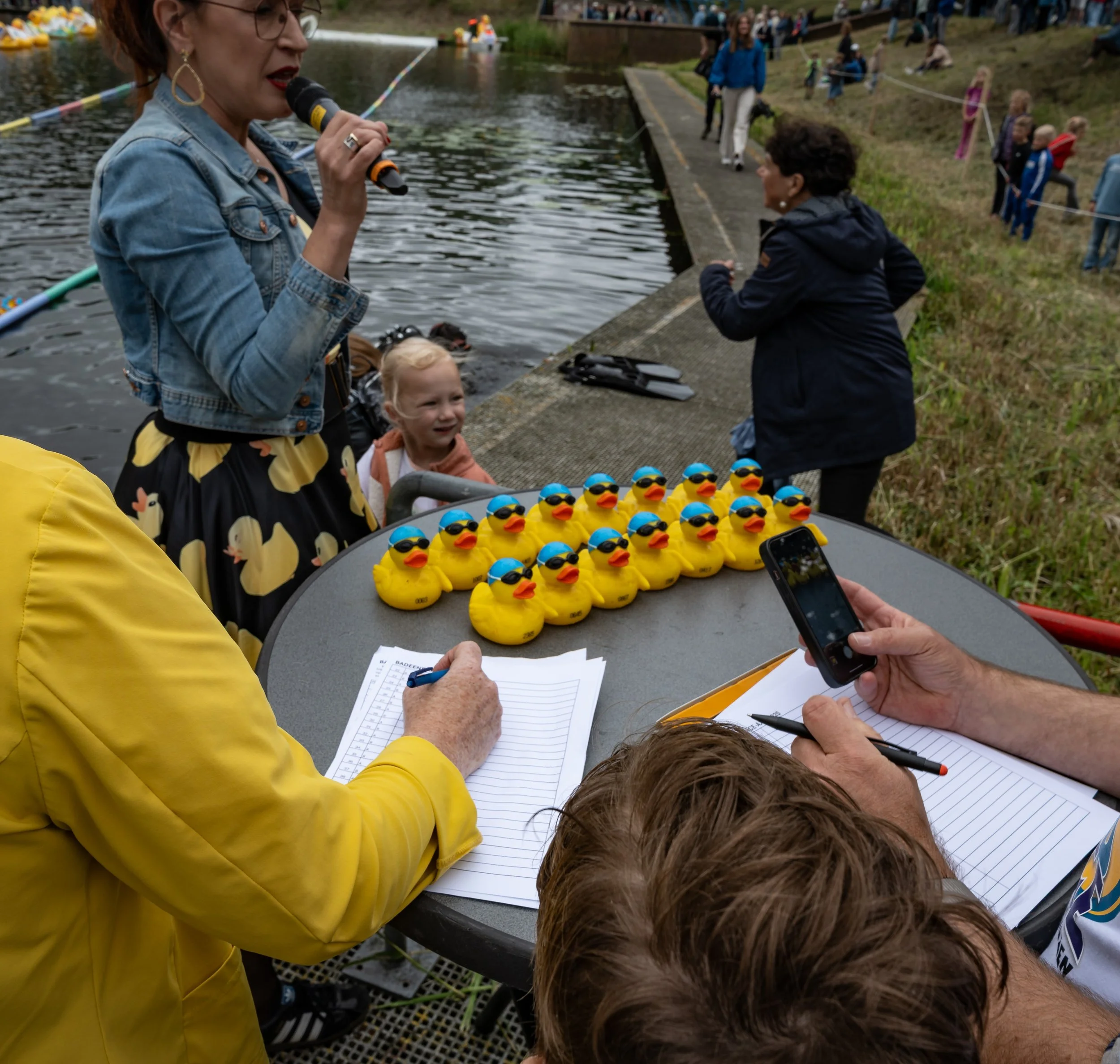 Nederlandse kindershow met rubber eendjes met zonnebrillen bij een rivier, met mensen die toekijken, en kinderen in de achtergrond op de oever.