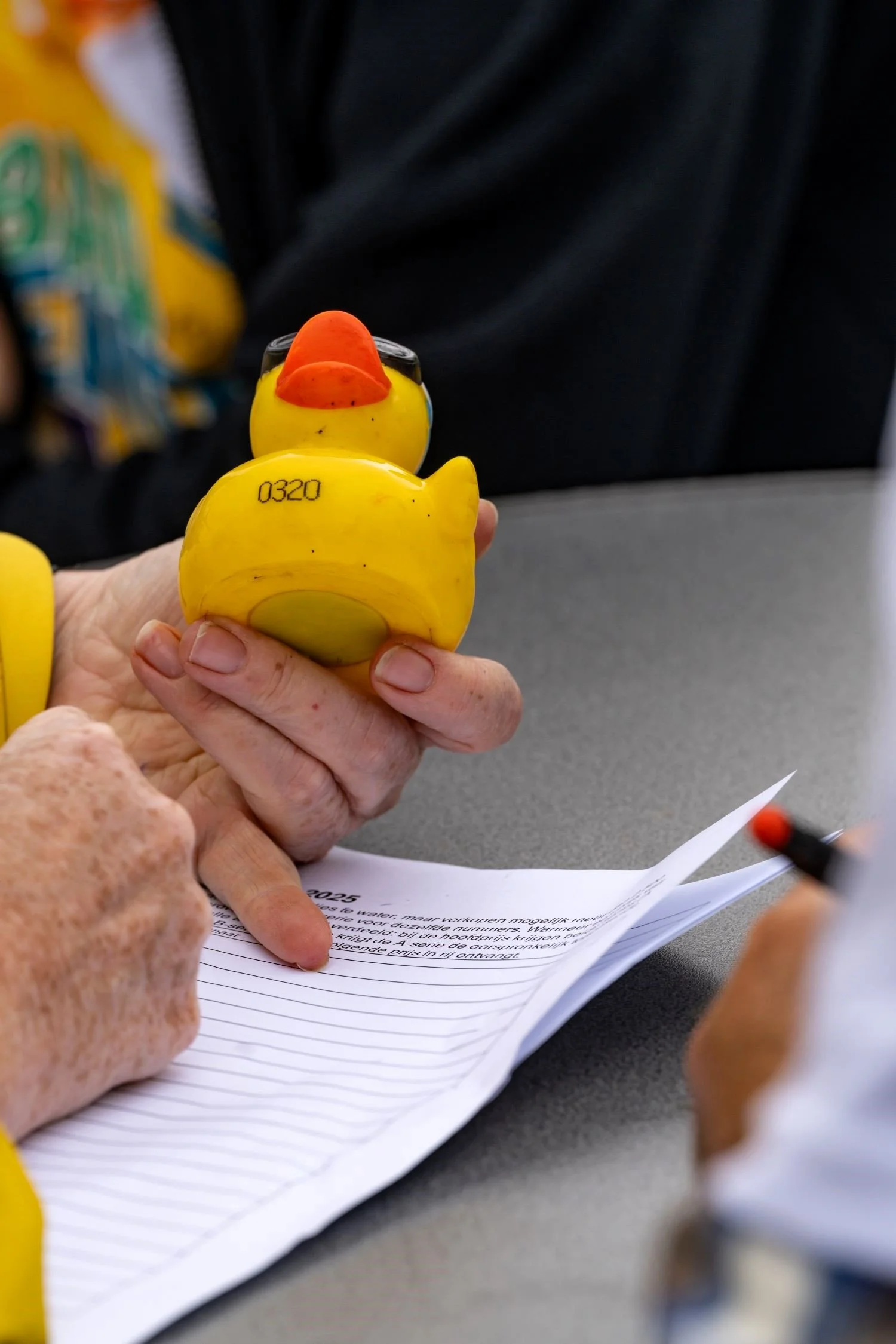Een hand houdt een rubberen rubber eend in de vorm van een gele vogel met een oranje snavel en zwarte ogen, terwijl iemand anders aantekeningen maakt op papier.