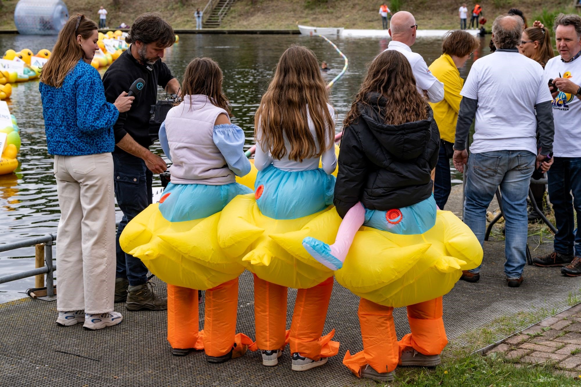 Mensen die zich verkleed hebben als eenden, met gele eendendekens en oranje benen, zitten aan de waterkant tijdens een evenement. Sommige mensen kijken naar het water, anderen staan erbij.
