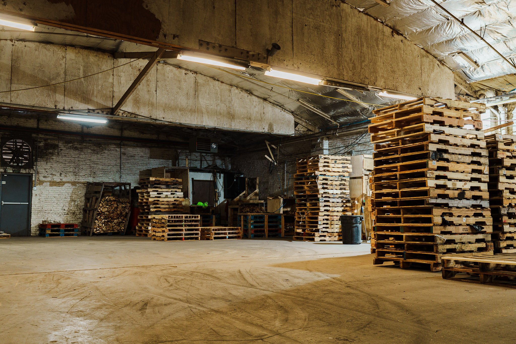 An industrial warehouse with a concrete floor and brick walls. Multiple stacks of wooden pallets are arranged on the right and against the back wall. Overhead fluorescent lights illuminate the space, which contains some tools and construction materials.