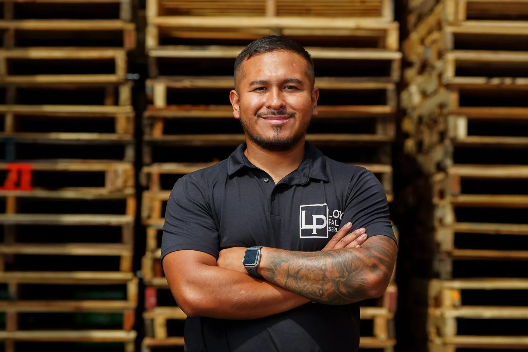 A man with short dark hair, a goatee, and a tattooed arm, standing confidently with crossed arms in front of stacked wooden pallets. He is wearing a black polo shirt with a logo on the chest and a smartwatch on his left wrist.