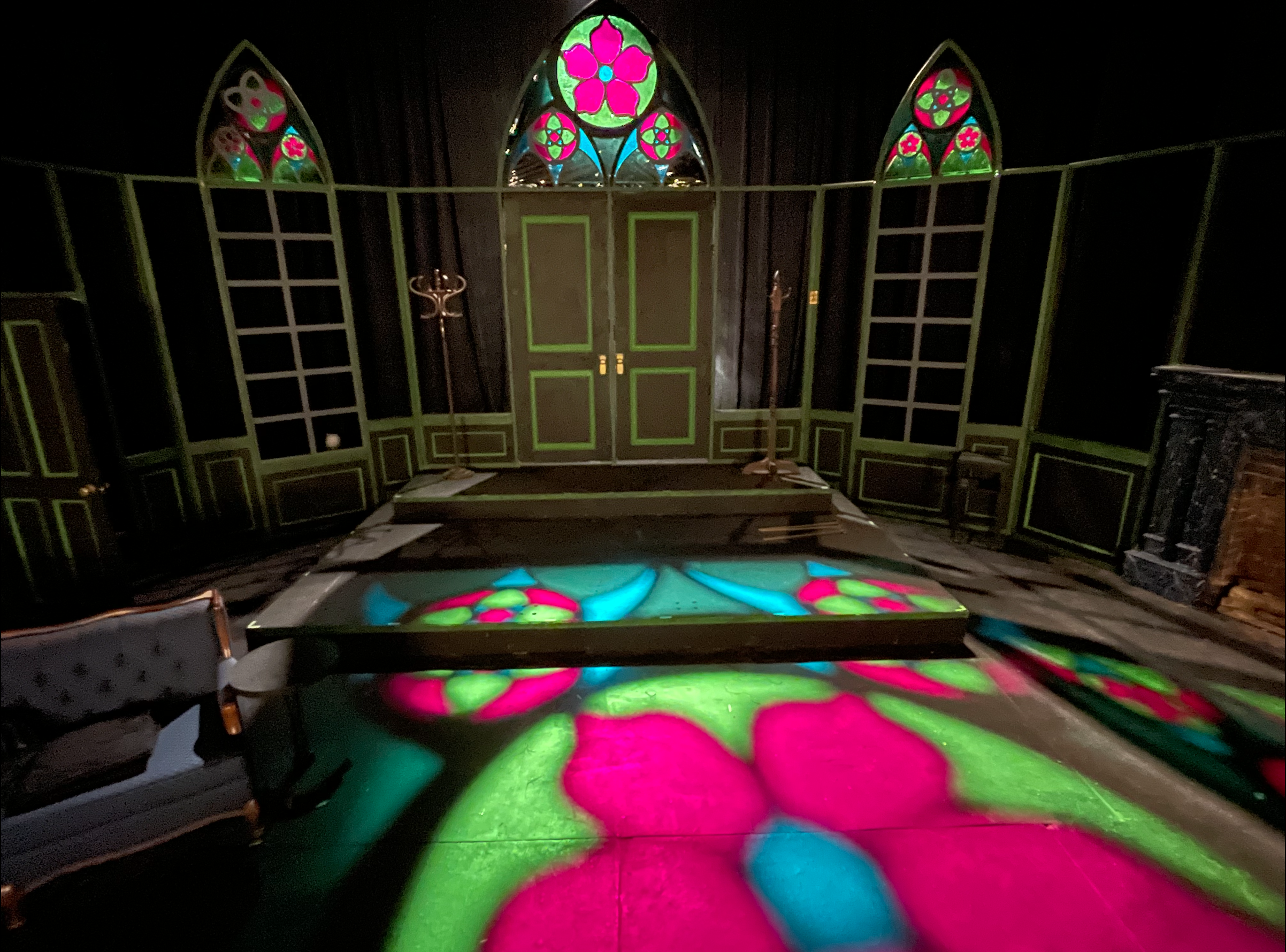 An empty dark room with stained glass windows projecting colorful pink, blue, and green light onto the floor and walls, with classic wooden furniture and two coat racks.