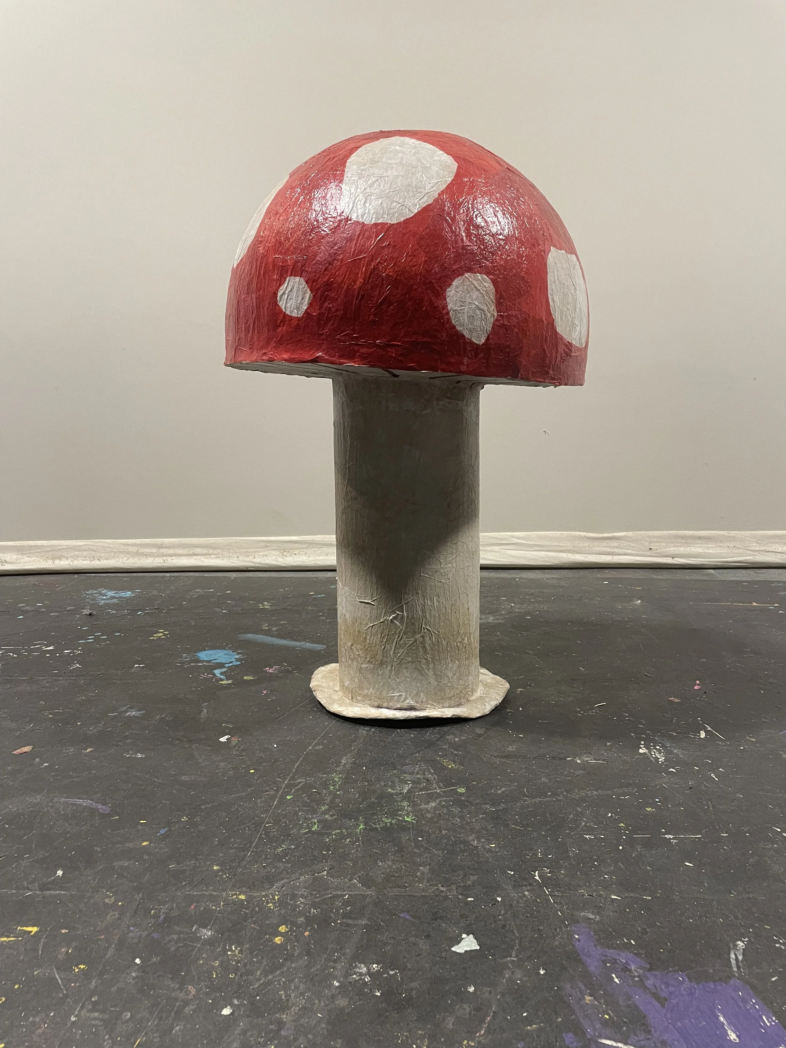A handmade model of a red mushroom with white spots, made of paper mache or similar material, standing on a black surface with paint splatters against a plain wall.