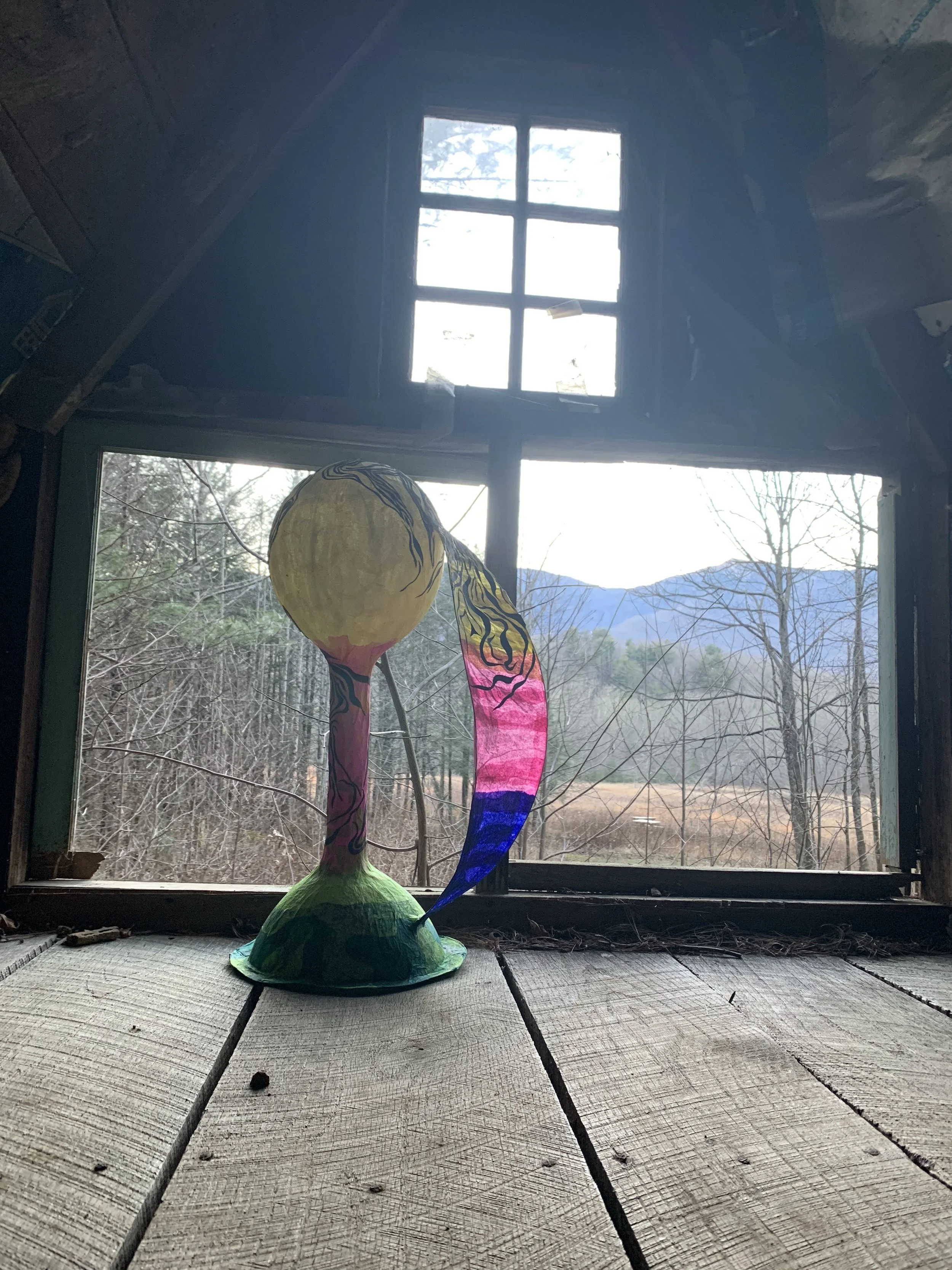 Colorful paper lantern with a landscape scene, placed on a wooden surface inside an attic, with a view of trees and mountains through the window.
