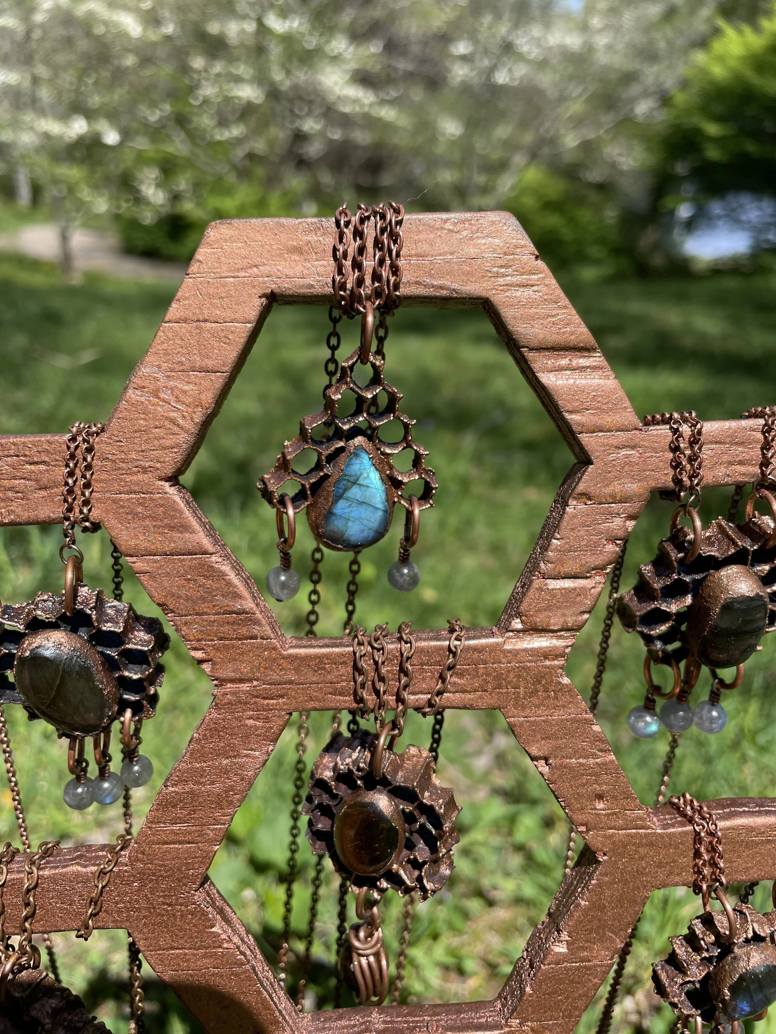A wooden jewelry display with hanging necklaces, featuring turquoise and dark stone pendants, set outdoors with green grass and trees in the background.