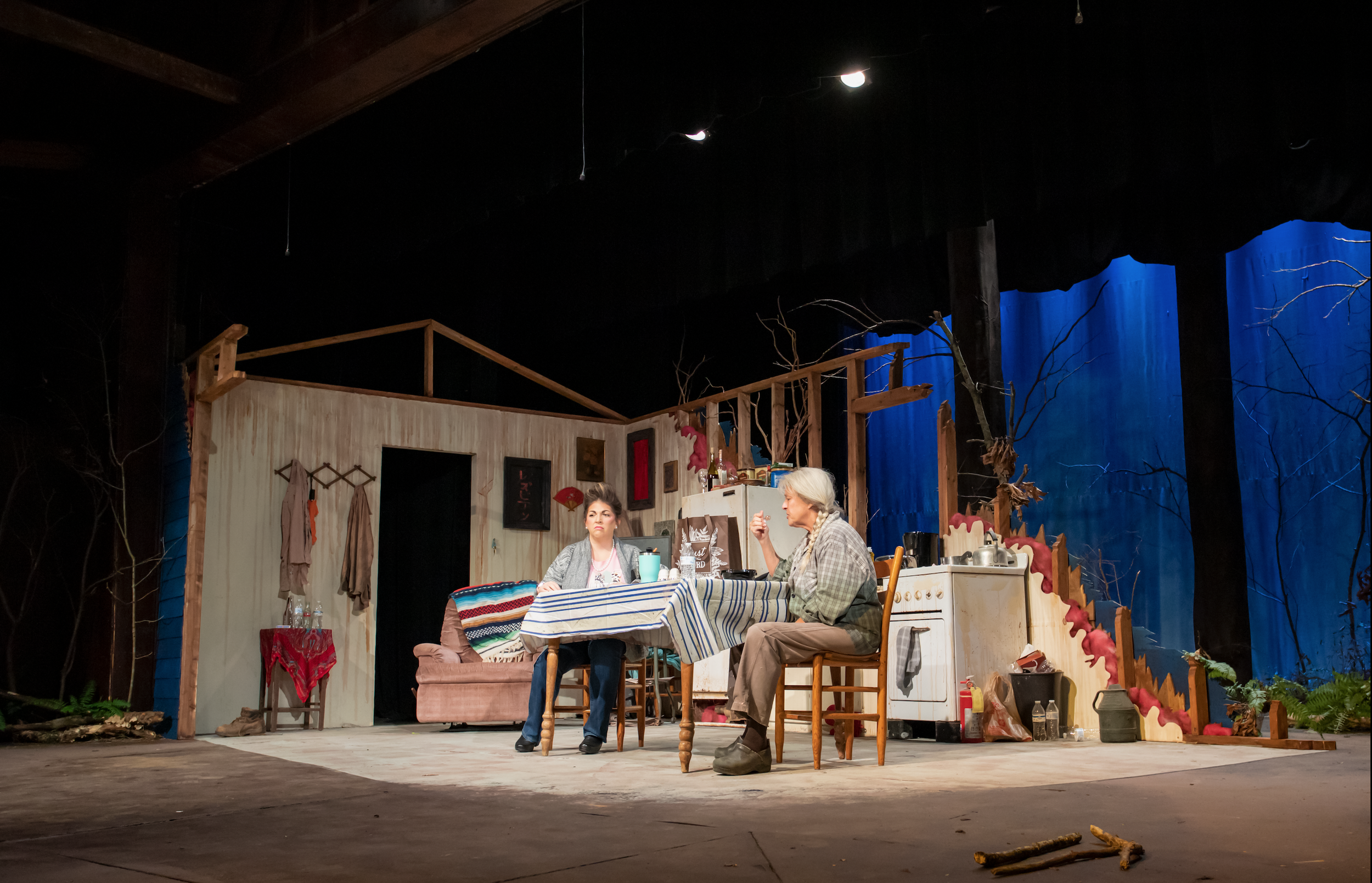 The image shows a stage set for a theatrical play depicting a rustic kitchen and dining area with two women seated at a table. The background includes a partial view of an interior room with wooden walls and hanging clothes, along with a staircase-li