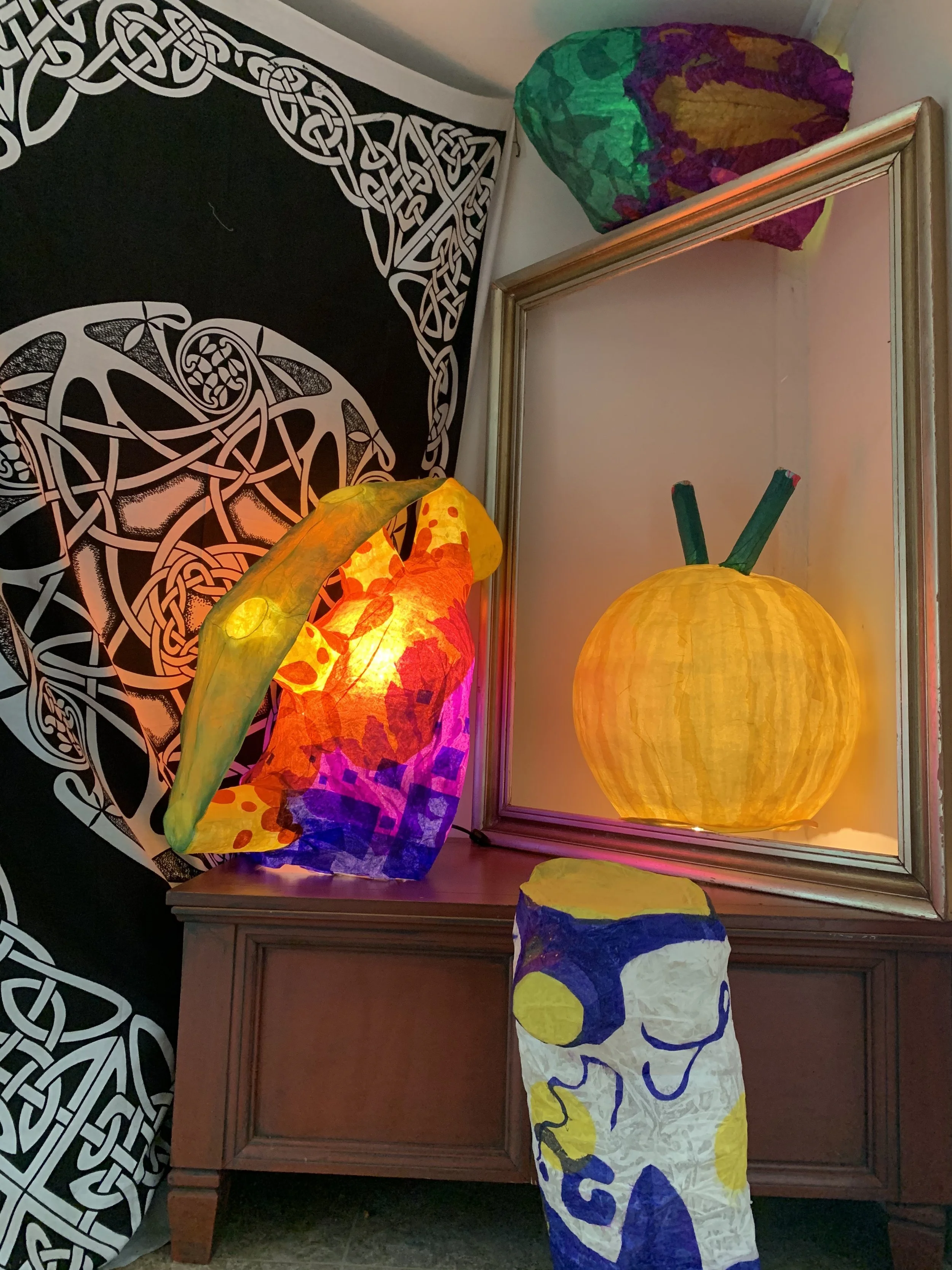 Decorative lanterns in various shapes and colors, including orange, yellow, purple, and green, placed on a wooden surface and inside a silver photo frame, with a black and white tapestry featuring Celtic knots in the background.
