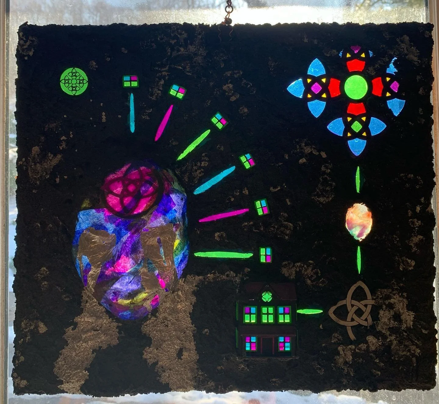 A stained glass artwork with a black background featuring a skull, a house, a cross, a circle, and abstract shapes in vibrant colors.