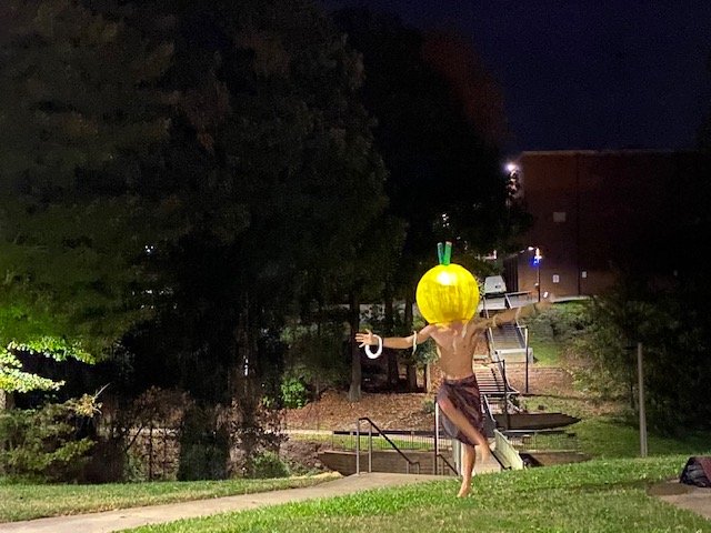 A person wearing a large yellow pumpkin head costume with green stem and leaf, standing outdoors at night, with arms outstretched and walking barefoot on grass.