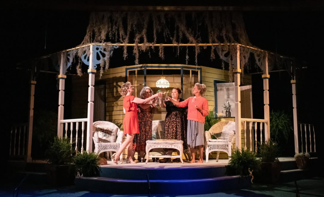 Five women standing on a covered porch raising glasses in a toast at night. The porch has a yellow house with green shutters and outdoor furniture. Hanging light illuminates the scene, and plants decorate the porch.