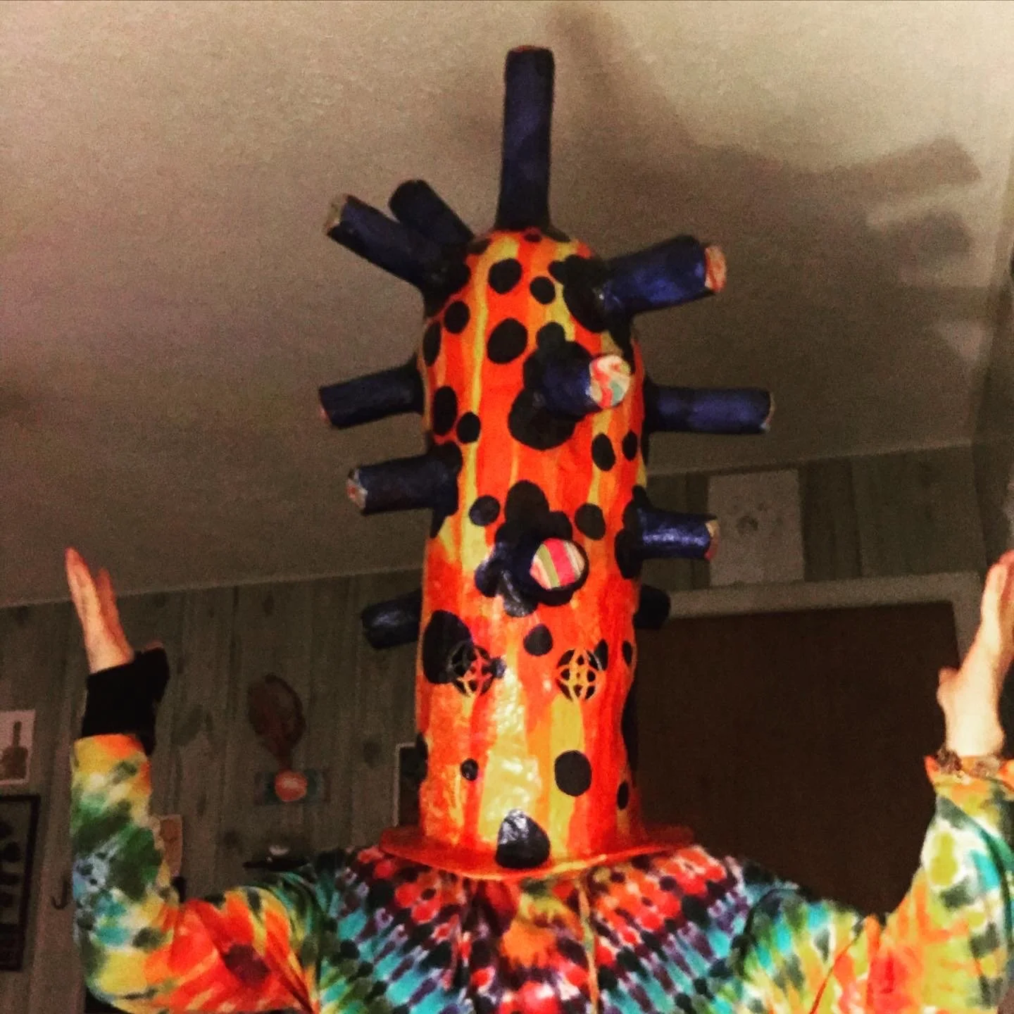 Person wearing a colorful costume with a large, painted giraffe head mask decorated with black spots and multiple dark blue horns, indoors with wooden-paneled walls.