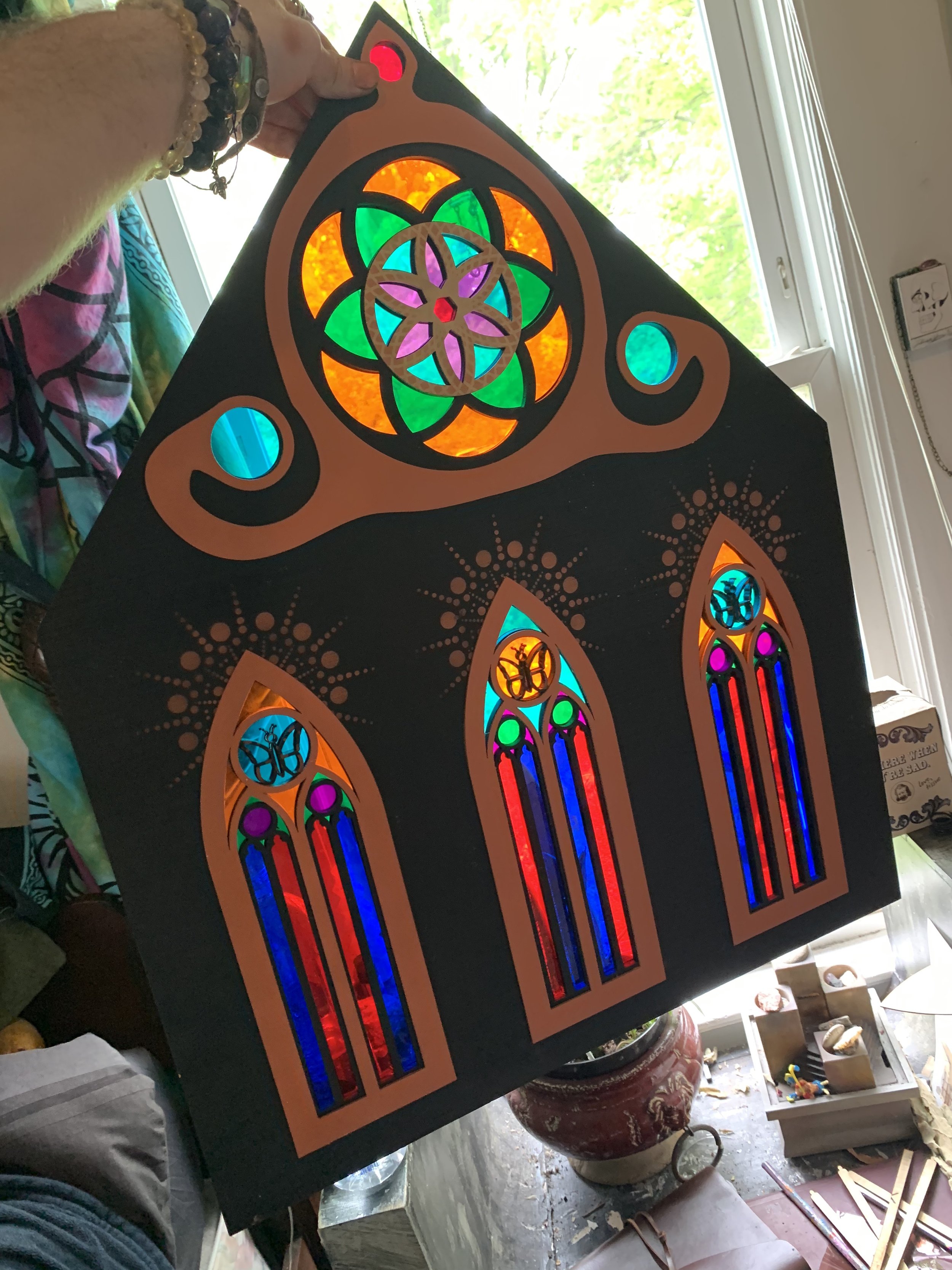 A person holding a colorful stained glass art piece with three tall, pointed arch windows and a circular stained glass design at the top, displaying vibrant colors like orange, green, blue, purple, red, and pink.