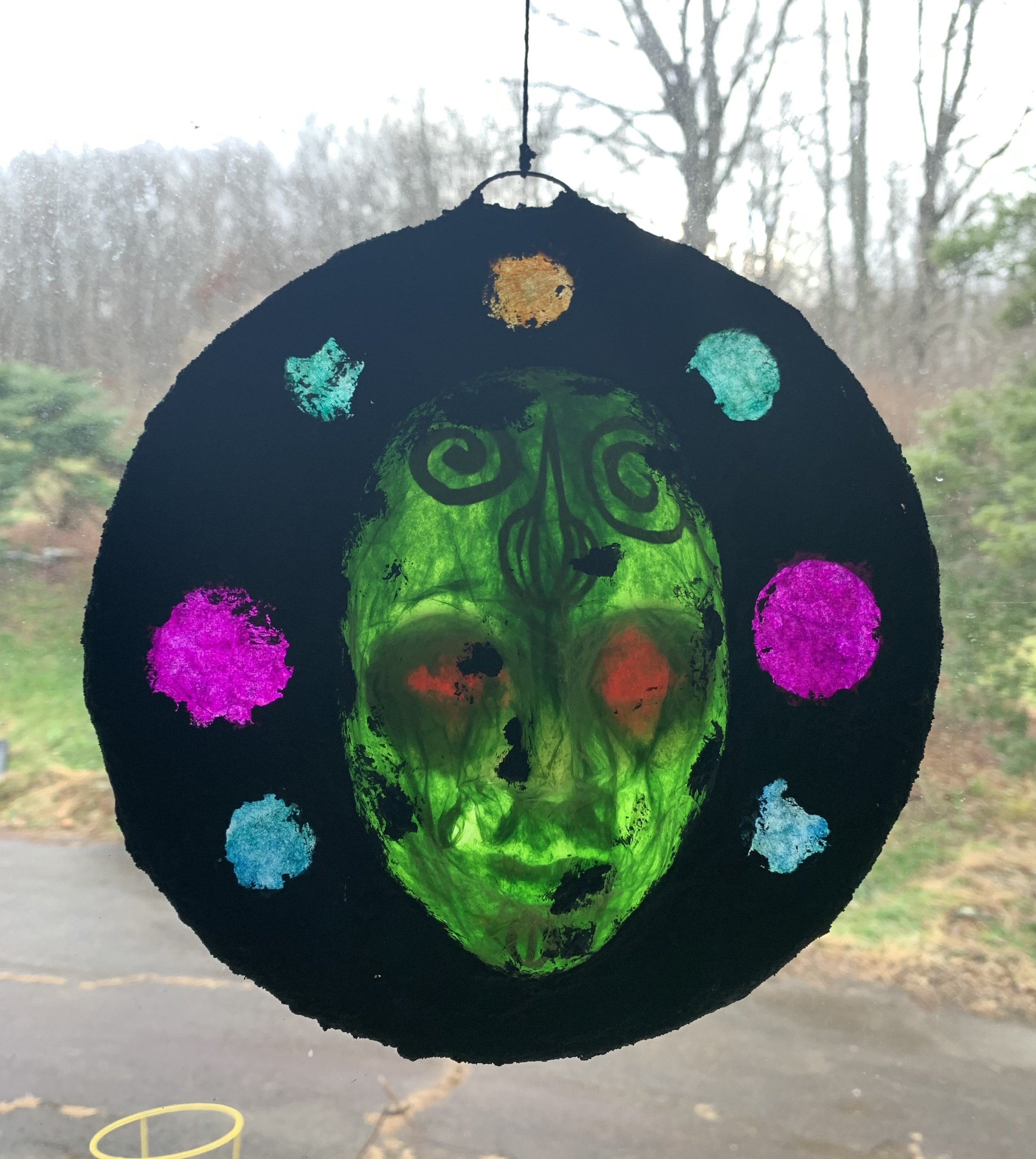 A decorative hanging lantern with a green skull face, surrounded by colorful dots, hanging in front of a window with trees outside.