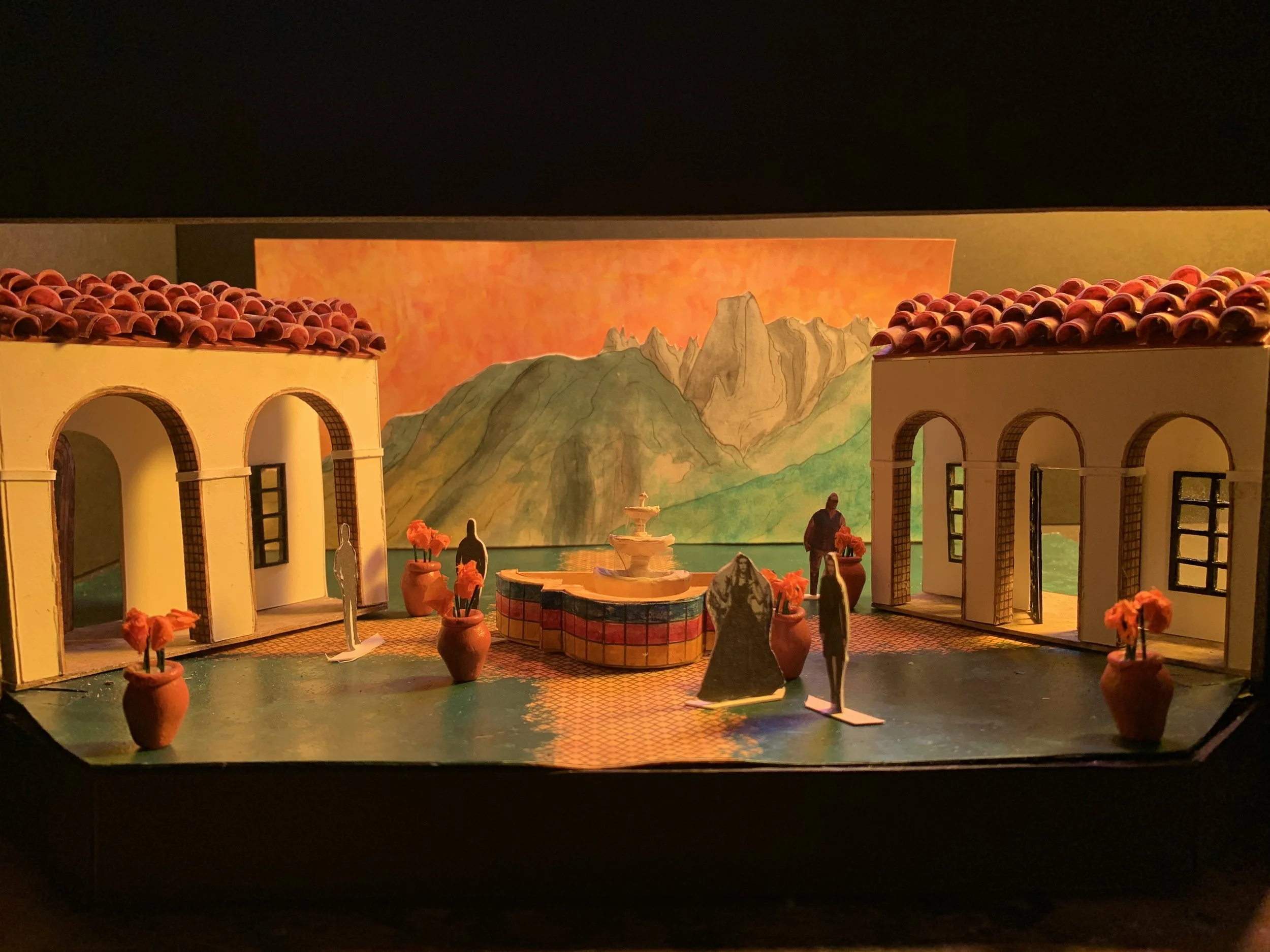 Miniature model of a courtyard with two yellow buildings with red tile roofs, a central fountain, and paper cutout figures of people. Background features painted mountains and sunset sky.