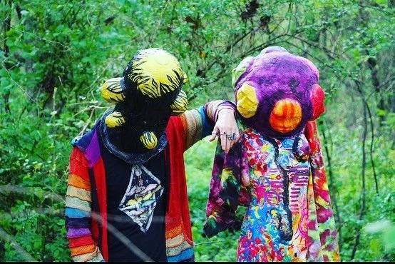 Two people wearing large, colorful mascot costumes standing in a green wooded area.
