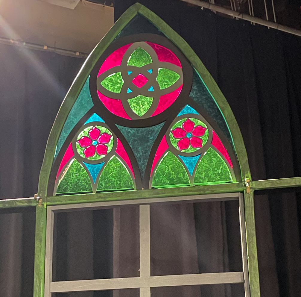 A stained glass window with geometric and floral designs in pink, green, and blue, set in a pointed arch frame.