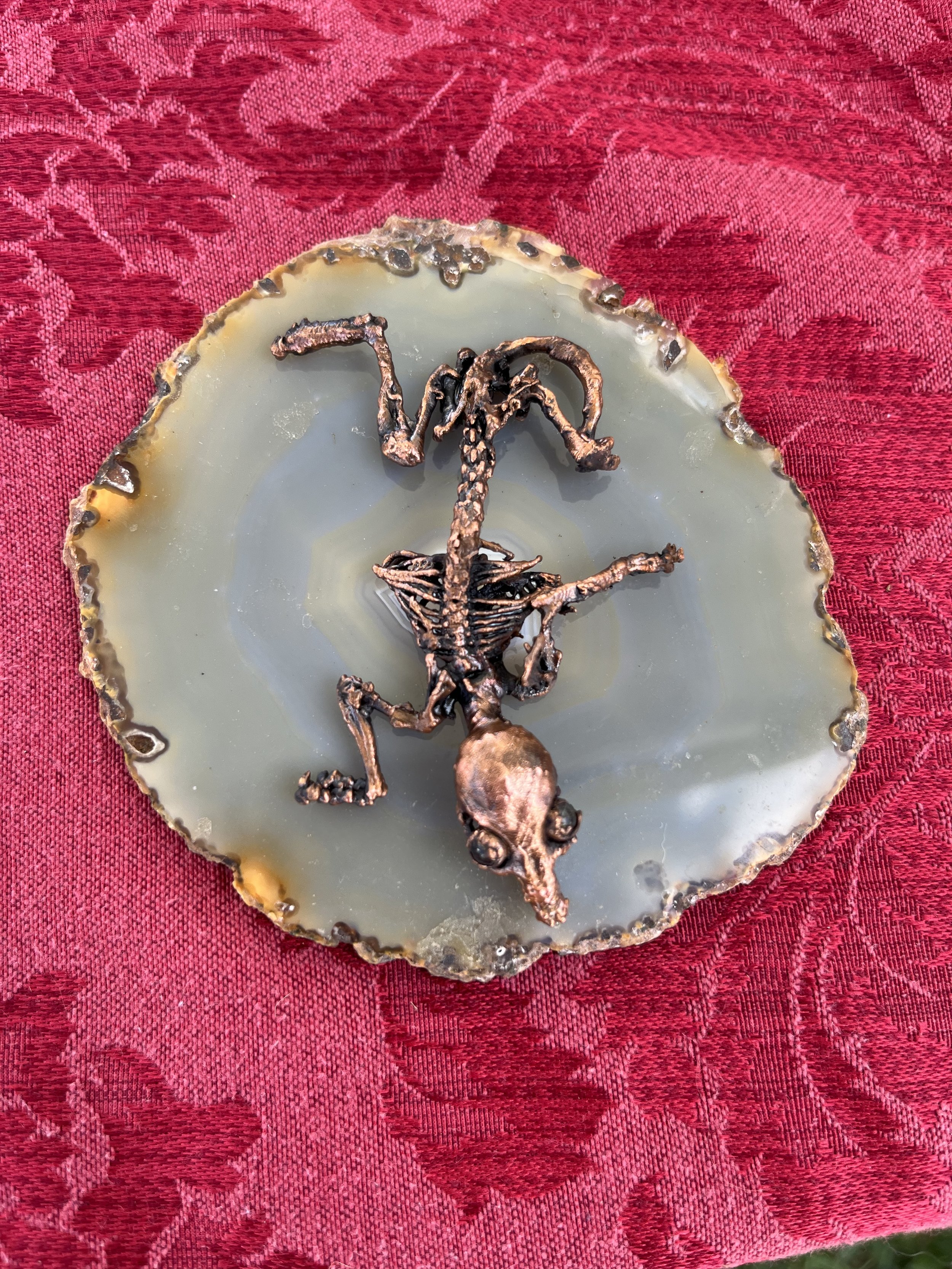 A small, detailed model of a dinosaur skeleton, likely a Tyrannosaurus rex, made of a metallic material, displayed on a polished, oval-shaped stone or mineral with a greenish hue, placed on a red patterned fabric tablecloth.