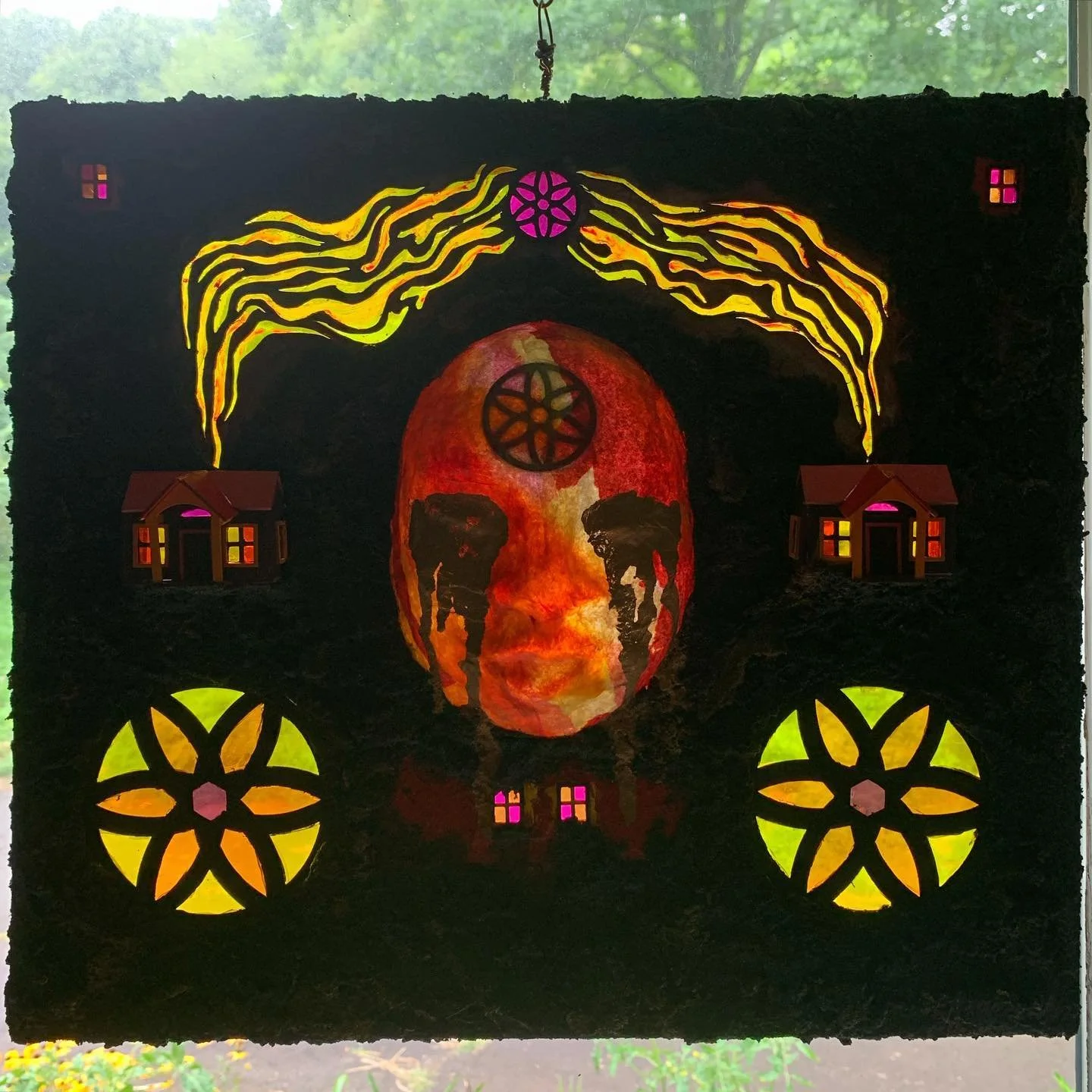 Art piece featuring a painted face with orange and red hues, surrounded by black, with yellow and pink geometric designs, small houses with lit windows, and stylized smoke or energy lines emanating from the top.