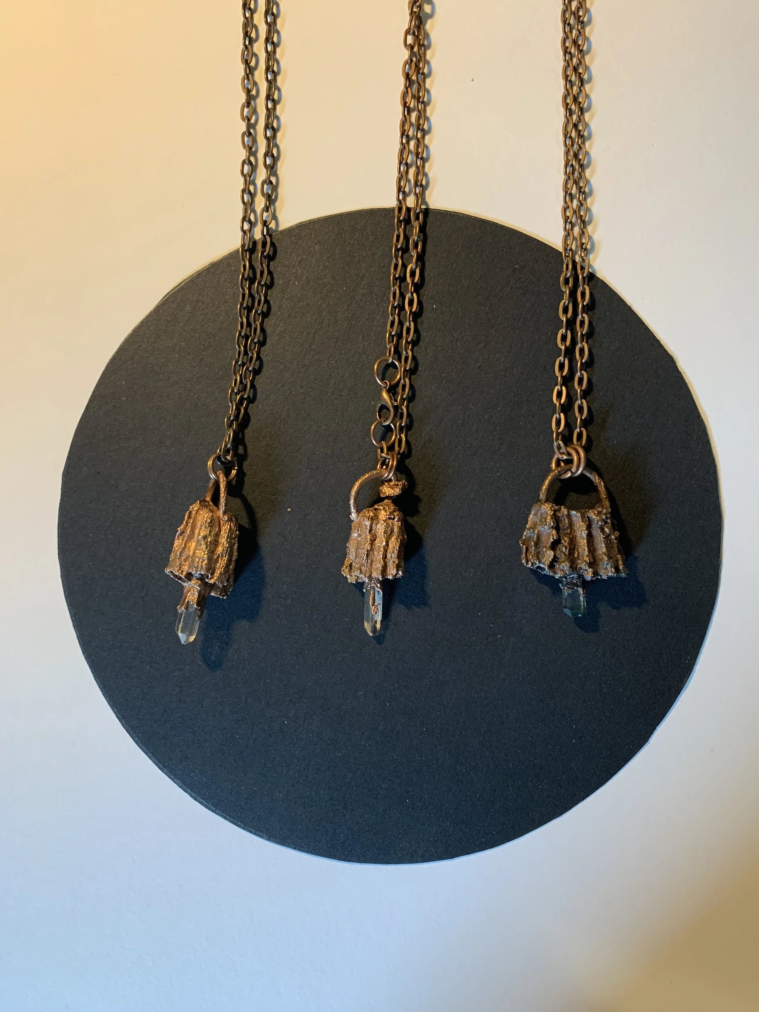 Three bronze necklaces with textured pendant charms, each adorned with a small crystal at the tip, hanging against a black circular background.