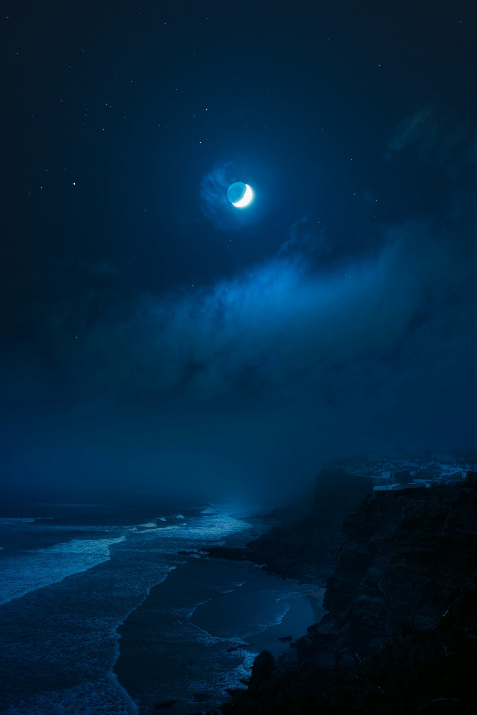 Nighttime view of a crescent moon over a rocky coastline with waves and mist, under a starry sky.