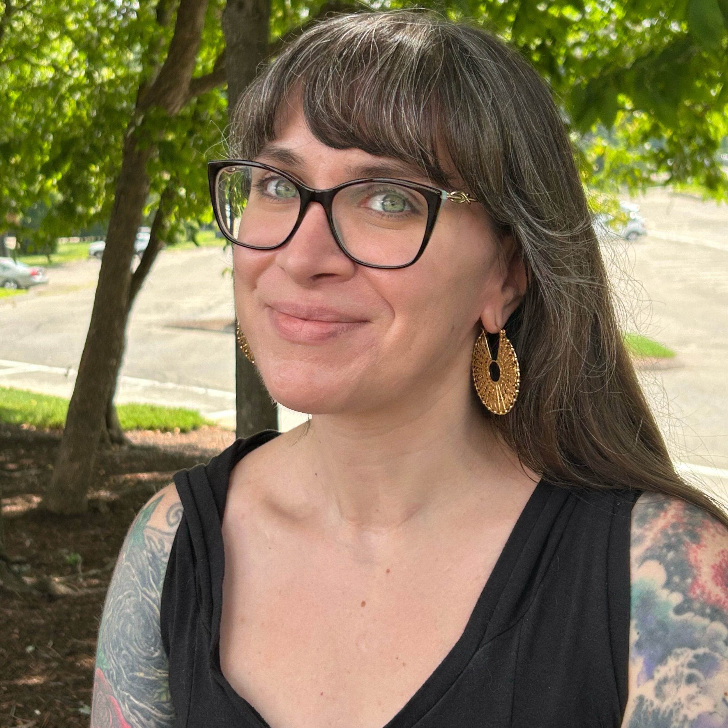 A woman with glasses and tattoos on her arms smiling outdoors, with green trees in the background.