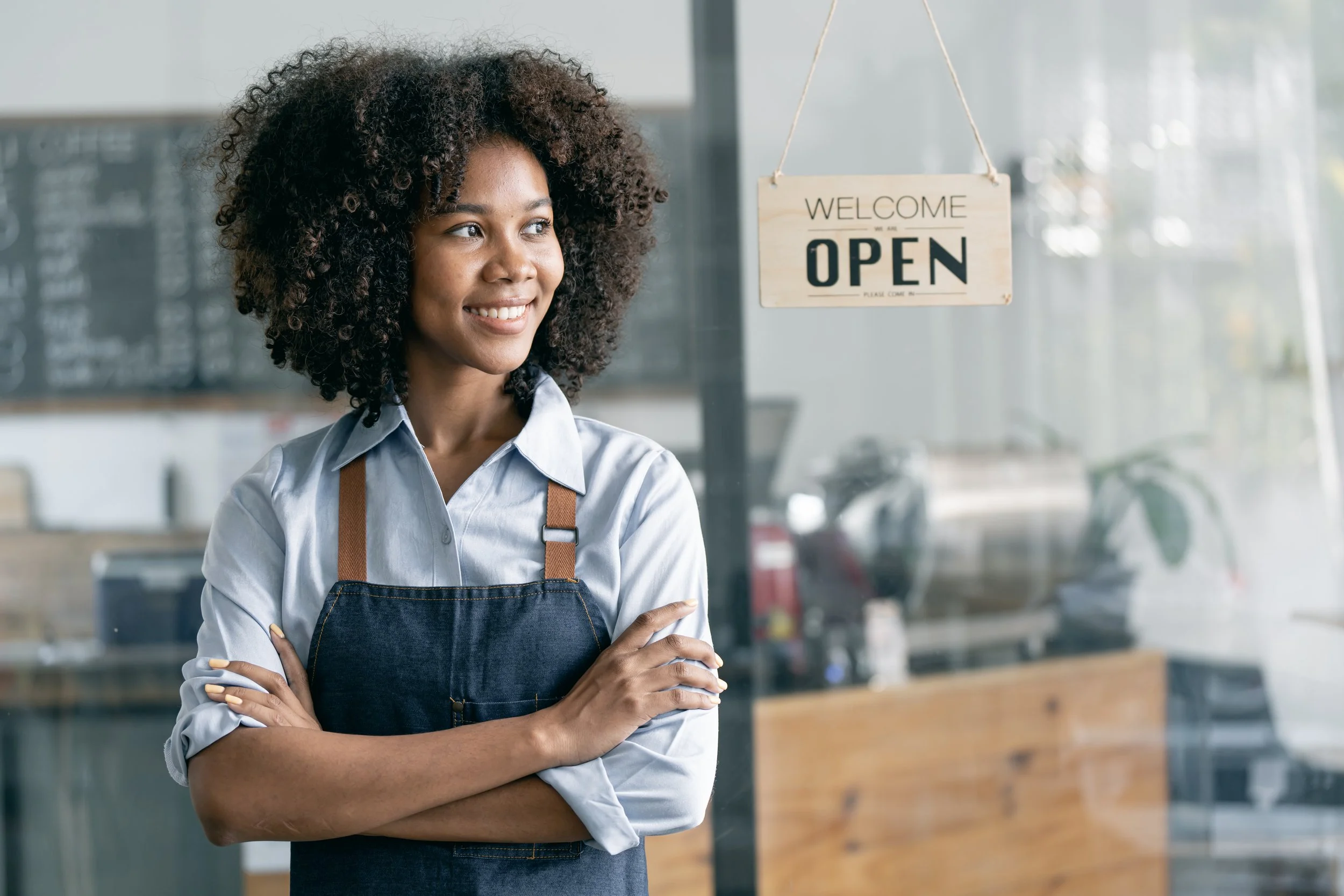 A smiling woman with curly hair wearing a light blue shirt and an apron standing inside a cafe with a welcoming smile, arms crossed, in front of a glass window with an "Open" sign