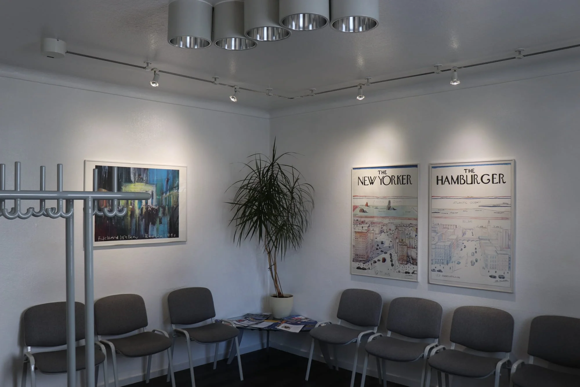 Wartezimmer mit weißen Wänden, mehreren grauen Stühlen, einem großen Pflanze, Wandkunst, darunter zwei Poster von The New Yorker und The Hamburger, und einer Deckenlampe.