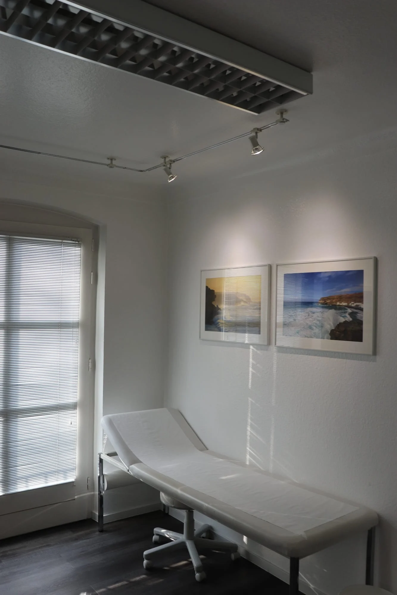 Behandlungszimmer mit Untersuchungsliege, Fenster mit geschlossenen Gardinen, zwei Fotografien an der Wand, Deckenleuchte, dunkler Boden.