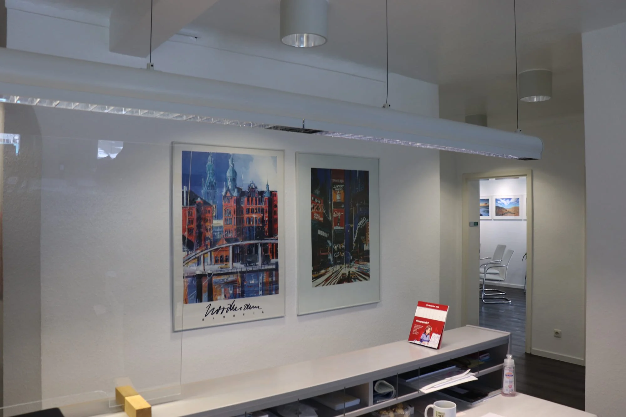 Ein Wartezimmer mit Wandbildern, einem weißen Schreibtisch, einem Handdesinfektionsmittel und einem Becher. Es gibt einen Durchgang zu einem Raum mit weißen Stühlen und Bildern an der Wand.
