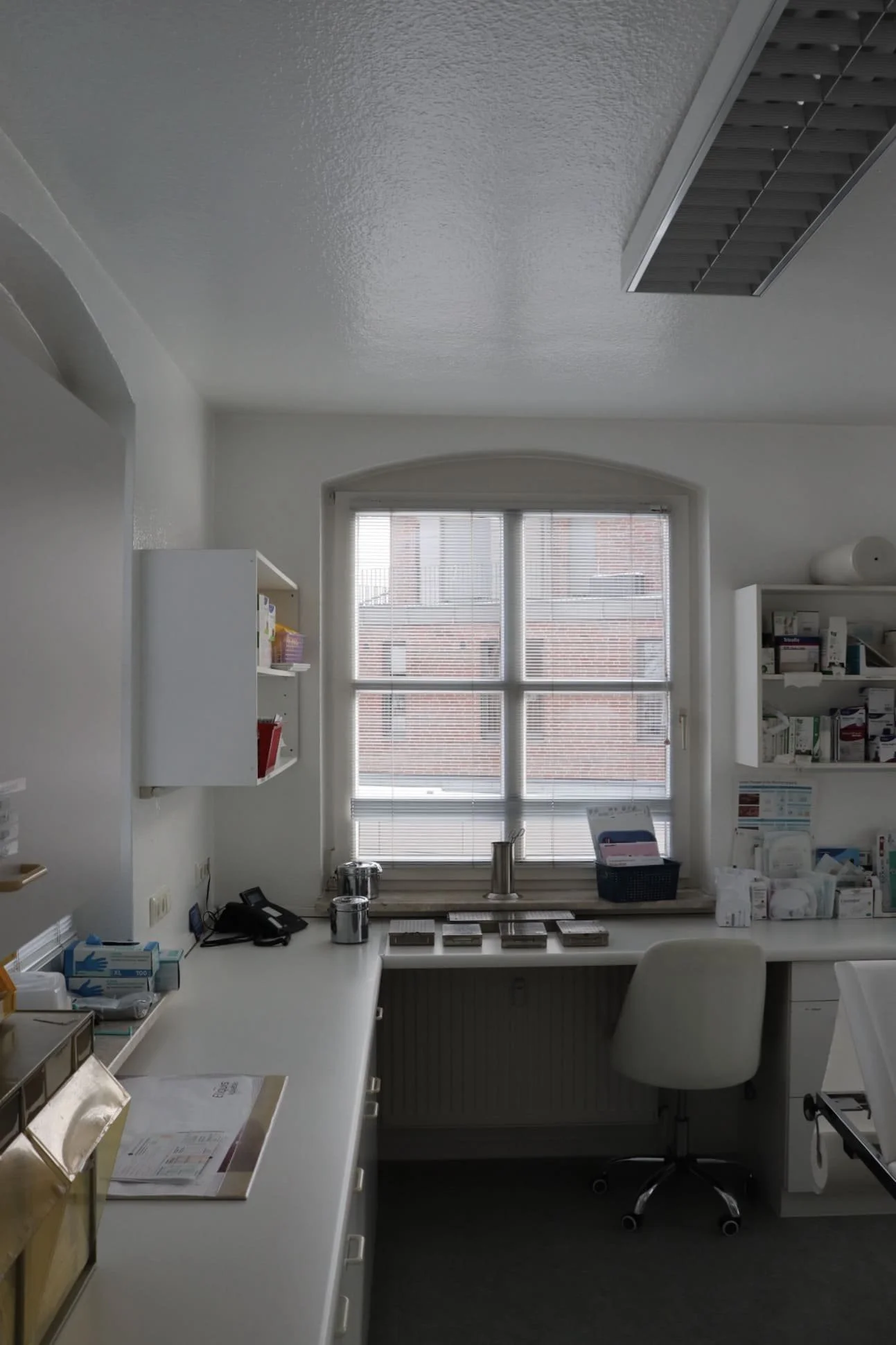 Ein heller Arzt- oder Praxisschreibtisch mit Stuhl vor einem Fenster, Regale mit medizinischen Produkten und Blumentöpfen auf der Fensterbank.