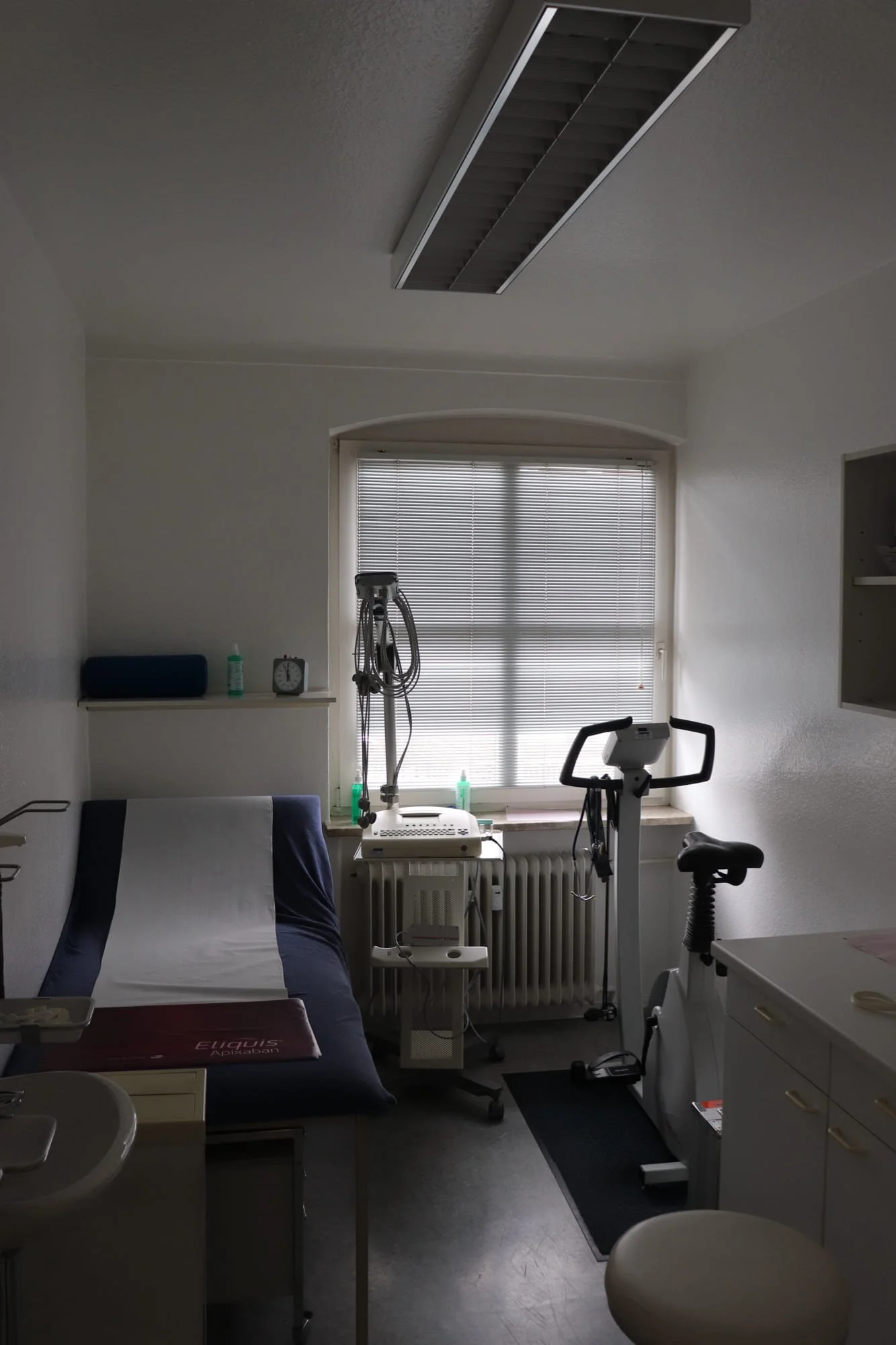 Blick in ein medizinisches Behandlungszimmer mit Bett, medizinischen Geräten und einem Fenster mit Jalousien.