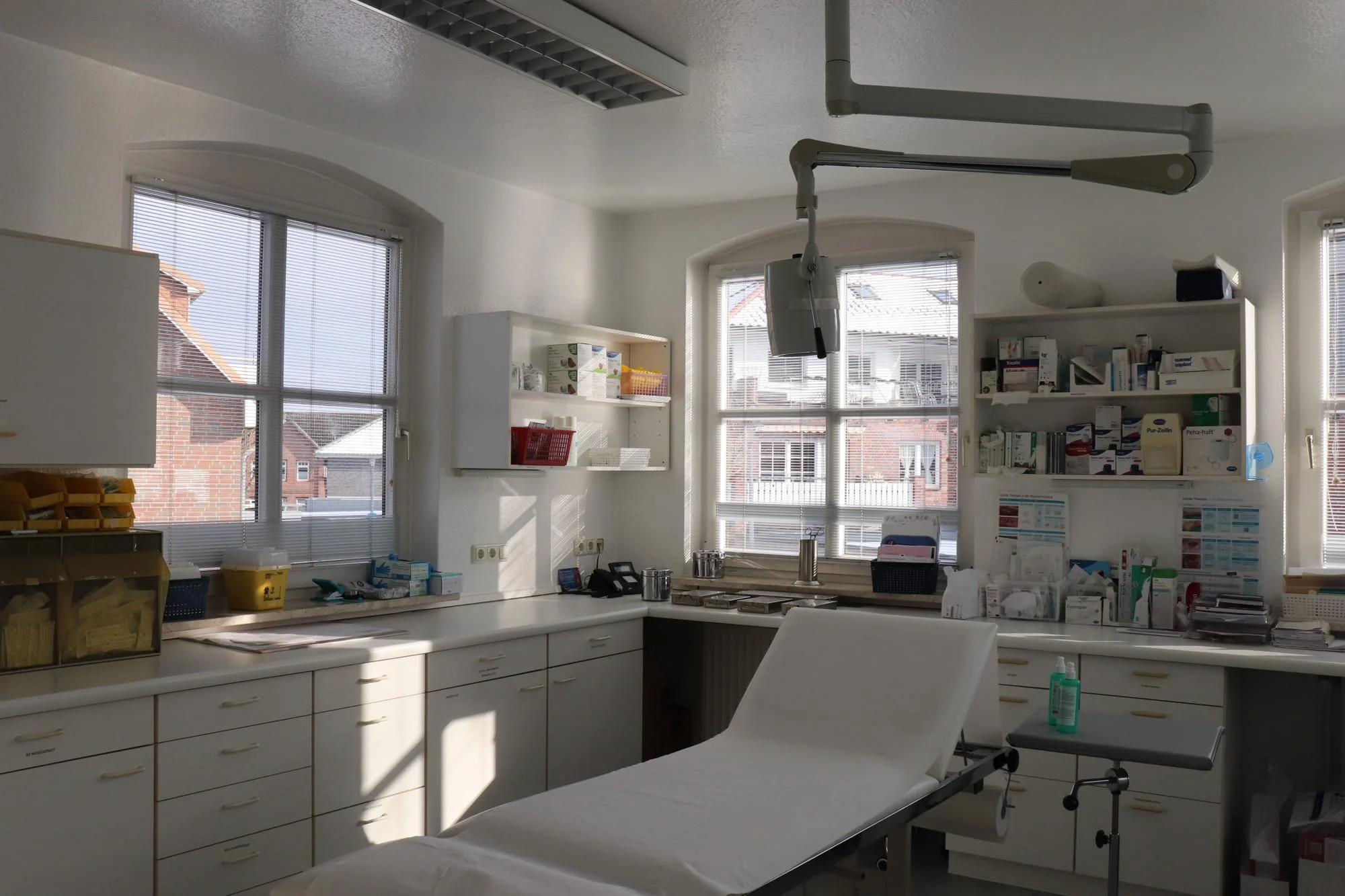 Ein leerer Behandlungsraum in einer Arztpraxis mit Untersuchungsliege, medizinischen Geräten und Medikamentenschränken, natürlichem Licht durch Fenster