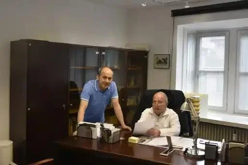 Zwei Männer im Büro, einer steht und lächelt, der andere sitzt am Schreibtisch mit Papieren in der Hand.