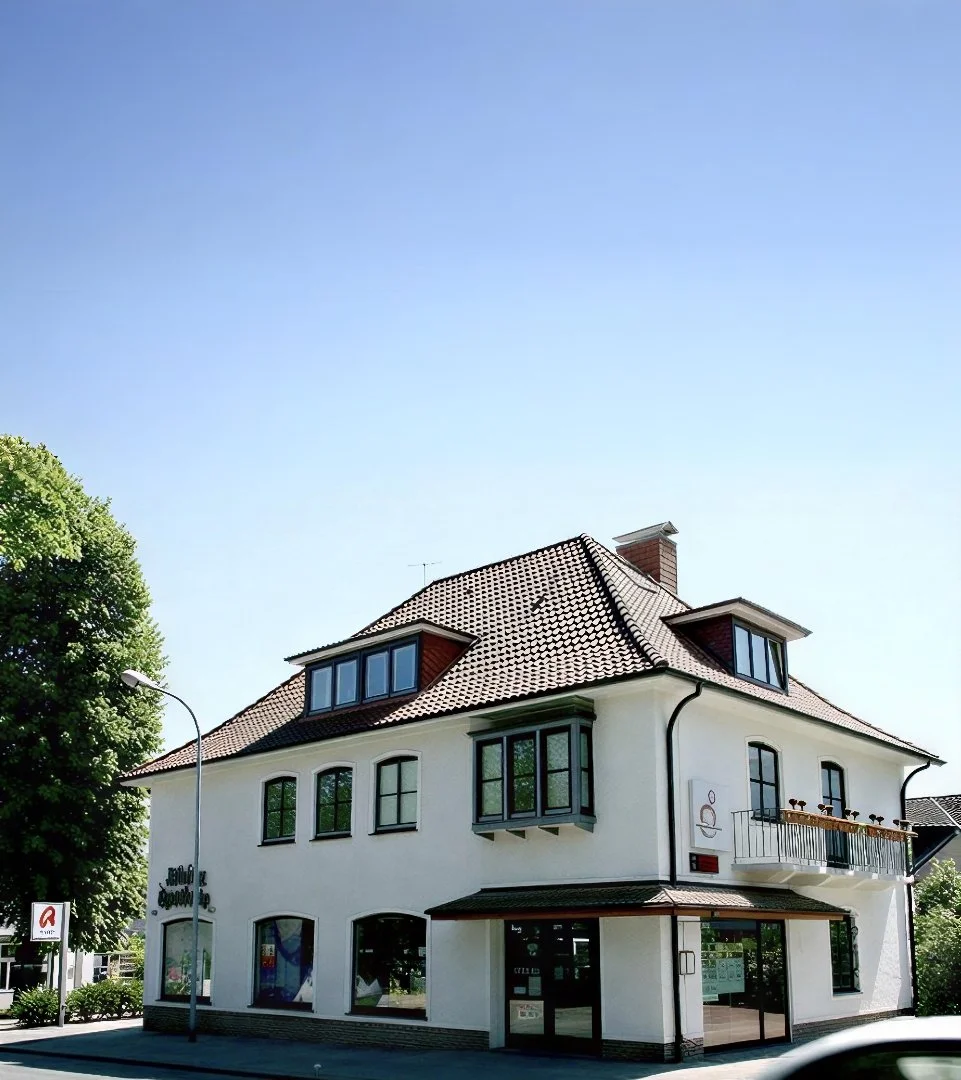 Ein weißes, mehrstöckiges Haus mit einem roten Ziegeldach, großen Fenstern und einem Balkon, umgeben von Bäumen, bei sonnigem Himmel.