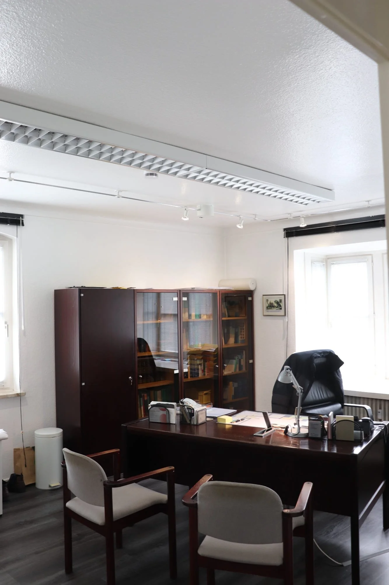 Büro mit dunklem Schreibtisch, schwarzen Lederstuhl, zwei weißen Stühlen, Büchern in einem Holzkabinet, Fenster mit Rollläden, hellgrauem Boden und weißen Wänden.