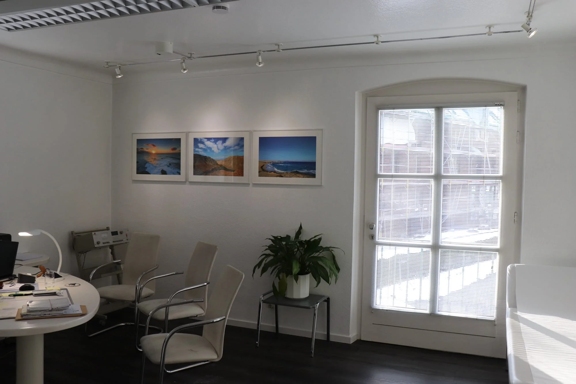 Innenraum eines Büros mit weißen Wänden, einem runden Tisch, sechs Stühlen, einem Fenster mit Rollos, einem Pflanzenkübel mit Pflanze, drei Bilder von Küstenlandschaften an der Wand, und einem hellen Sofa