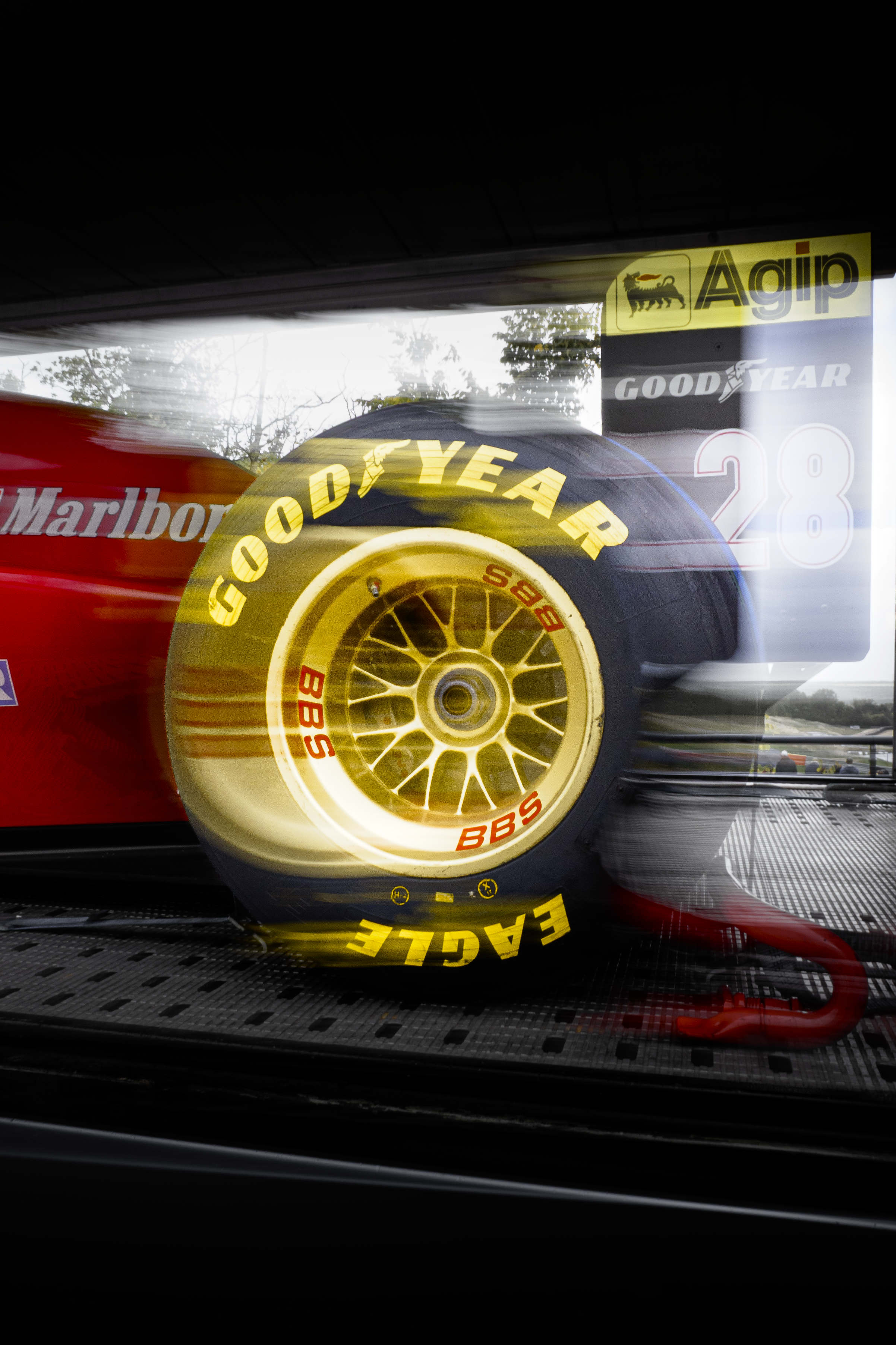 Une photo d'une voiture de course vue de côté, avec un focus sur la roue avant, qui porte des pneus Goodyear avec des inscriptions en jaune. On voit également des logos de sponsors comme Marlboro, Agip, Goodyear, et d'autres, sur la voiture.