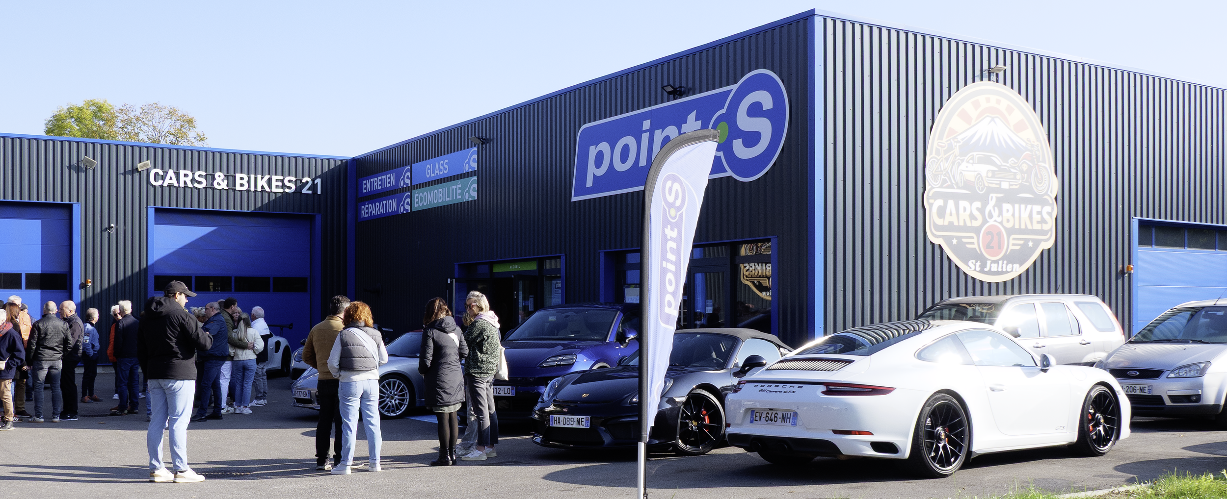 Une file de personnes attend devant un bâtiment bleu foncé avec un panneau 'CARS & BIKES 21'. Plusieurs voitures de luxe, y compris une Porsche blanche décapotable, sont garées devant le bâtiment. Il y a aussi un drapeau avec le logo 'point S'.