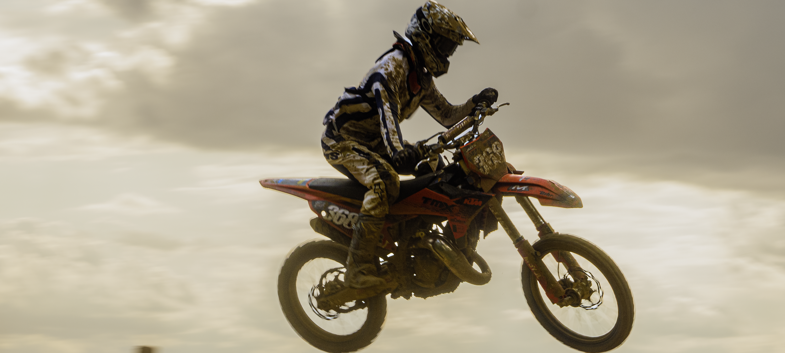 Coureur de motocross en pleine saut sur sa moto orange, silhouette contre un ciel nuageux