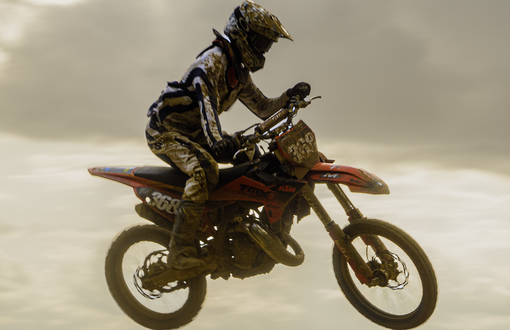 Motocross en l'air avec un pilote portant un casque et un équipement de protection, sur une moto orange dans un environnement poussiéreux.