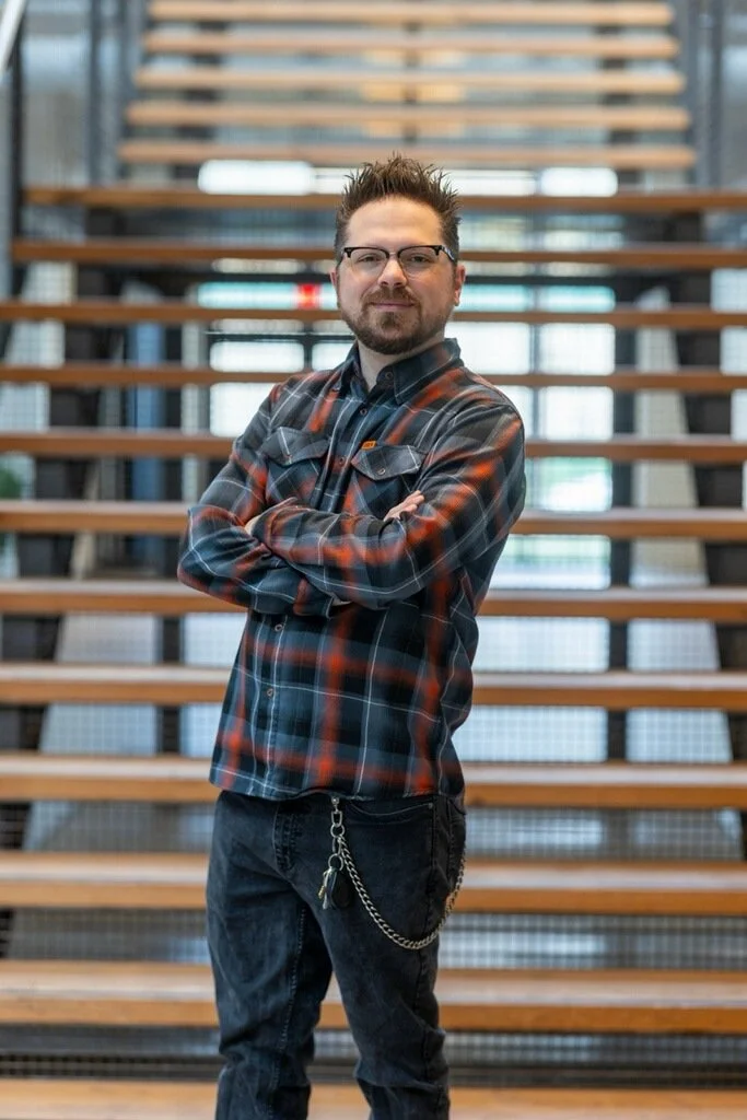 A man with glasses, a beard, and short spiked hair, wearing a plaid shirt and black jeans with a chain, stands confidently with his arms crossed in front of a wooden staircase in a modern building.