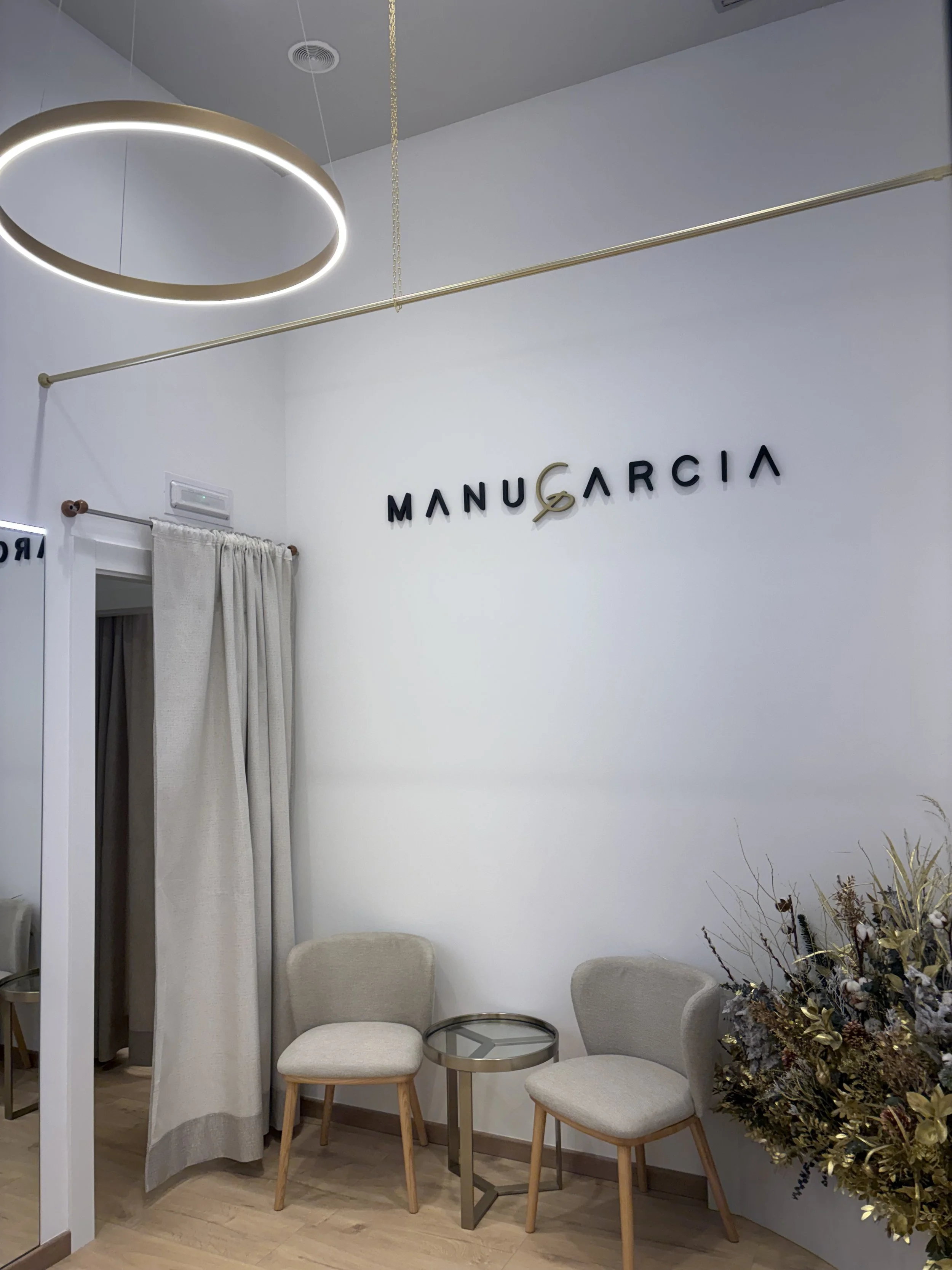 Interior de un consultorio o sala de espera con paredes blancas, con dos sillones beige, una mesa auxiliar de metal y vidrio, un ramo de flores artificiales o secas en una esquina y un letrero que dice 'Manu García' en la pared.