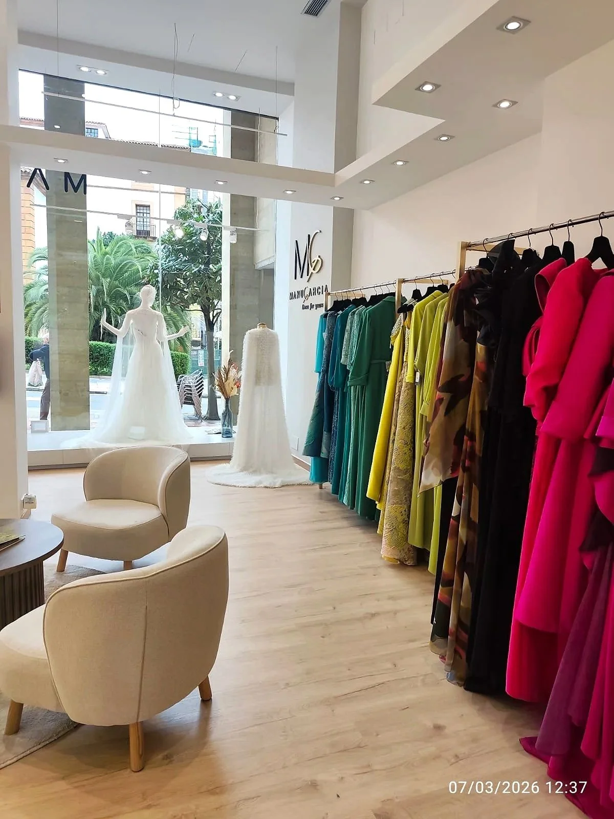 Vista del interior de una tienda de moda, con maniquíes con vestidos de novia, estanterías con prendas de colores, y sillas cómodas en un ambiente moderno y luminoso.