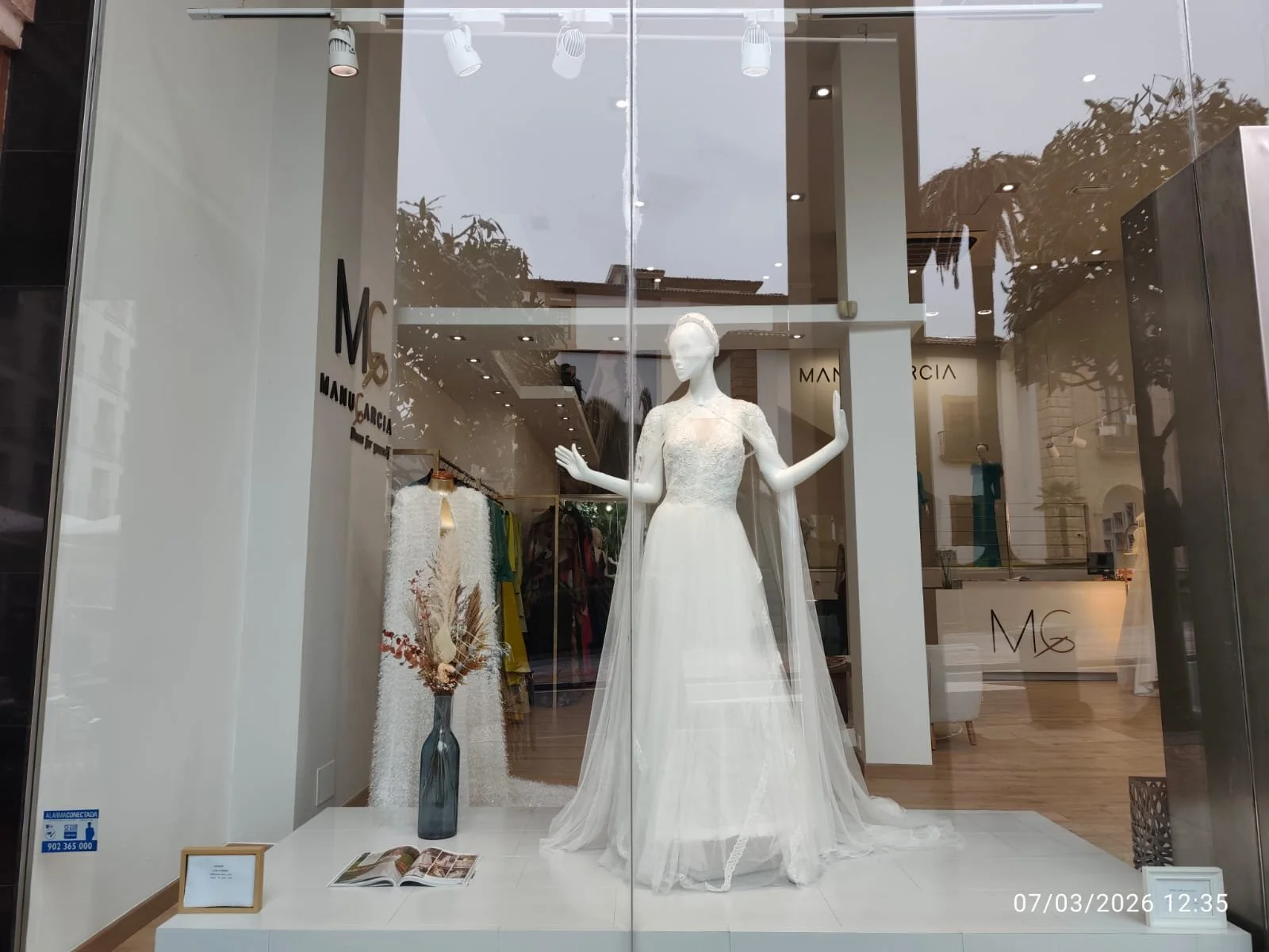 Vitrina de una tienda de ropa con un maniquí vestido con un vestido de novia. Hay una pared blanca, un letrero que dice M&G Manuela García, y un ramo de flores en un florero en la parte inferior izquierda. La tienda tiene exhibiciones de ropa en el fondo.