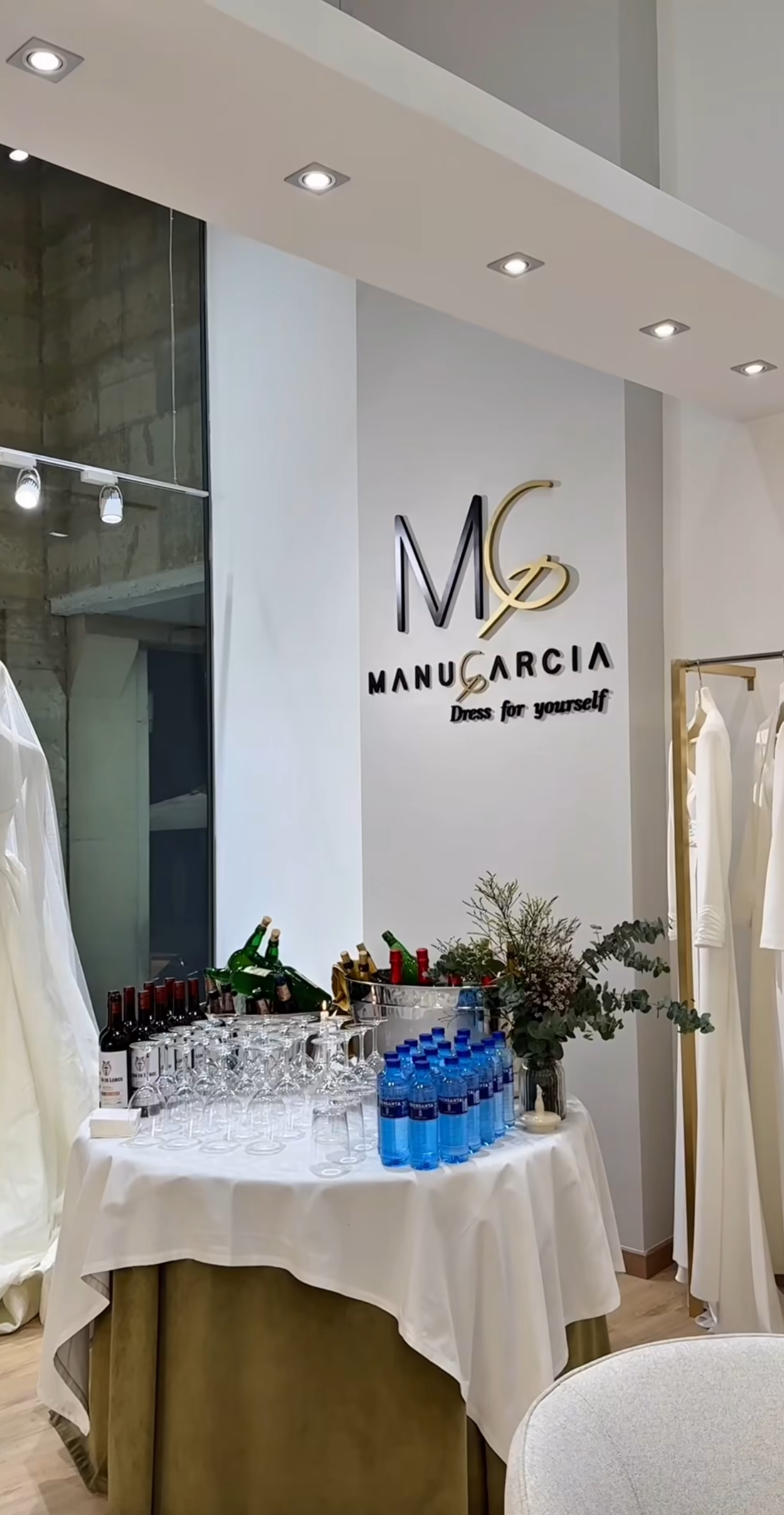 Mesa decorada con copas, botellas de agua y de bebidas alcohólicas, floreros y plantas ornamentales en un showroom de moda llamado "Manu García" con logo y lema "Dress for yourself" en la pared de fondo.