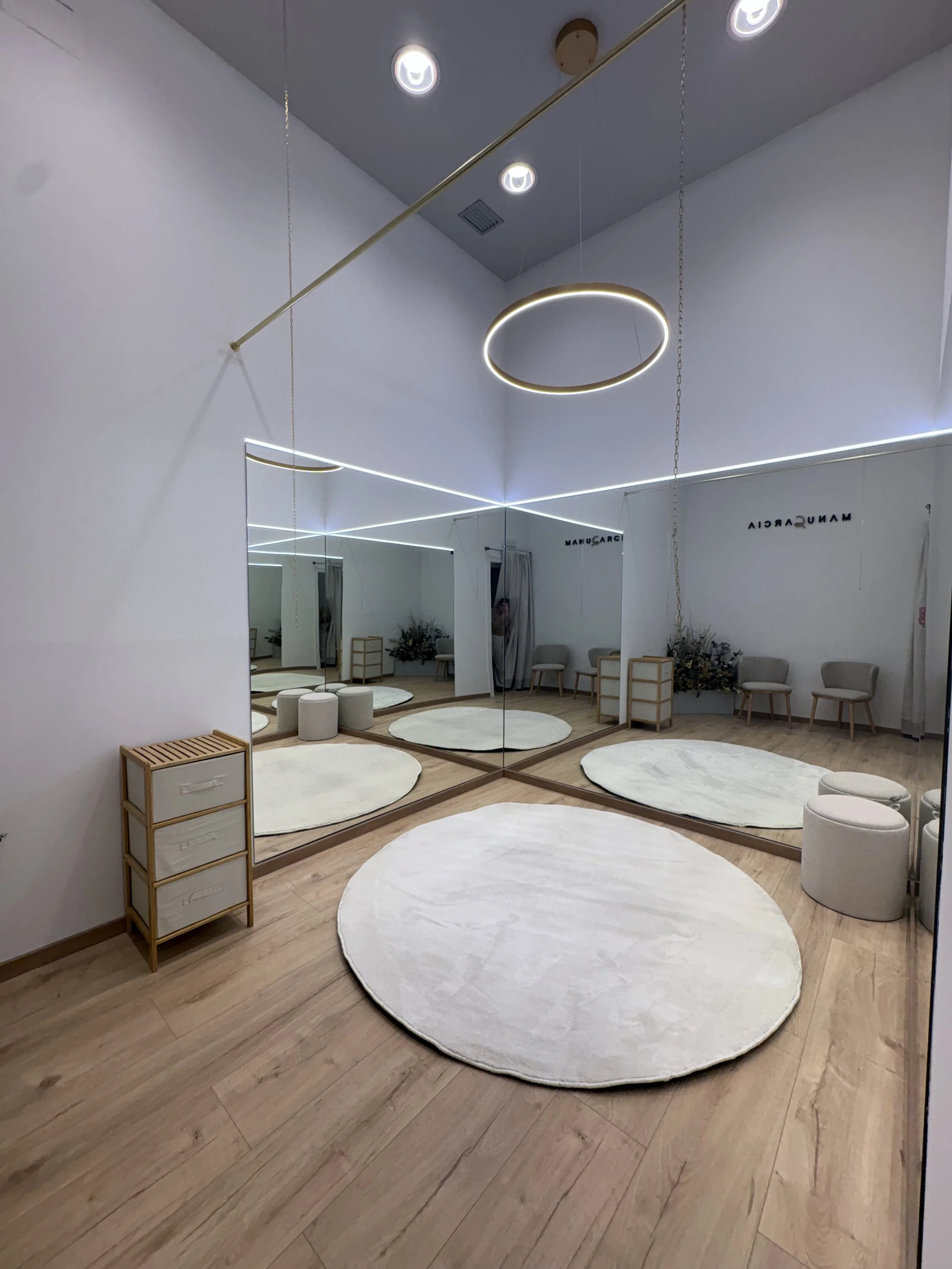 Sala de espera con paredes blancas, suelo de madera clara, alfombra circular blanca, sillas grises, espejos grandes en una pared, y una lámpara de aro colgante con iluminación LED, todo en un diseño minimalista y moderno.