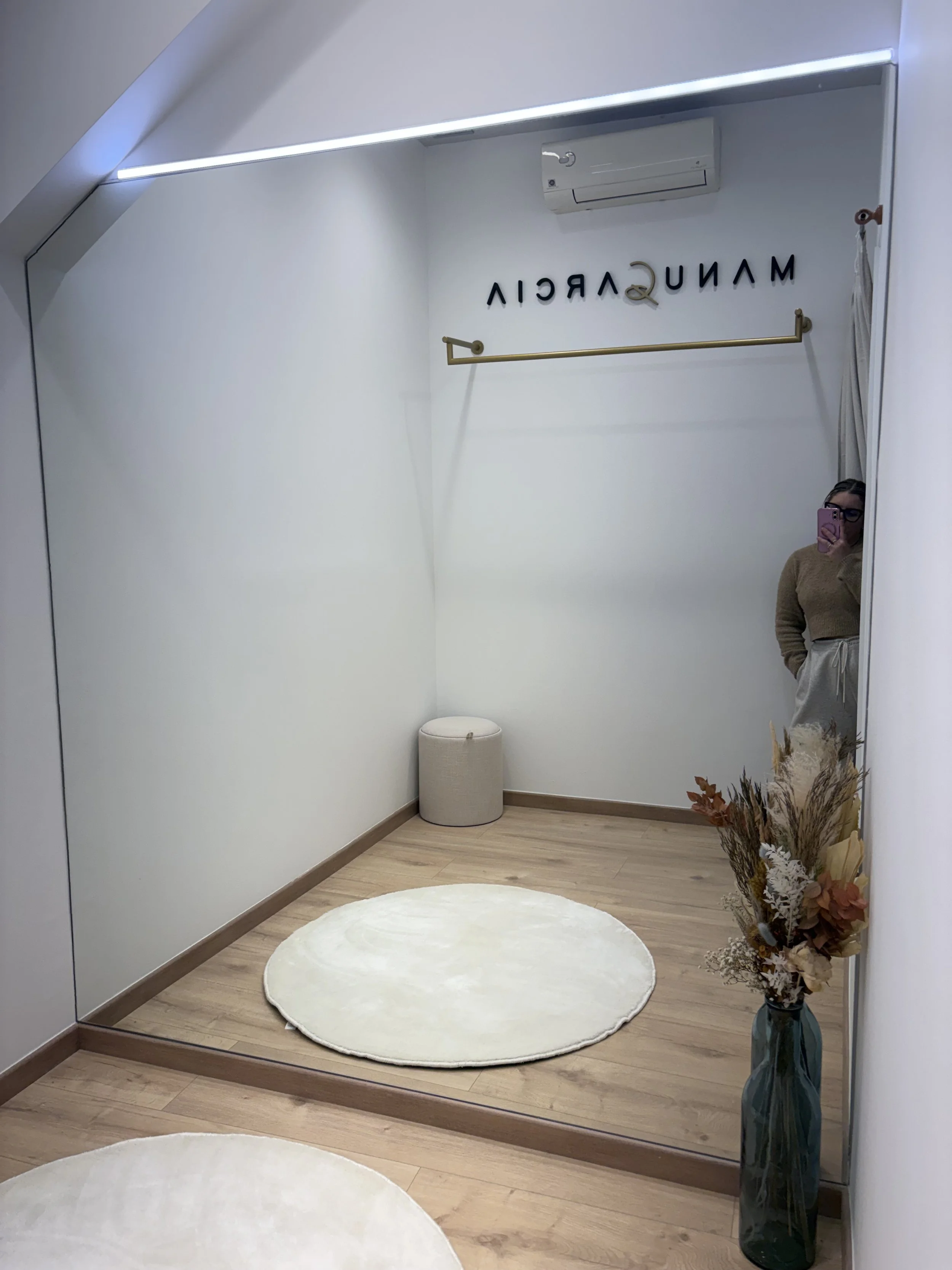 Vestidor minimalista con espejo grande, sillón beige, alfombra blanca redonda, vaso con flores secas en la esquina, y un letrero que dice 'MANIQUERÍA' en la pared.